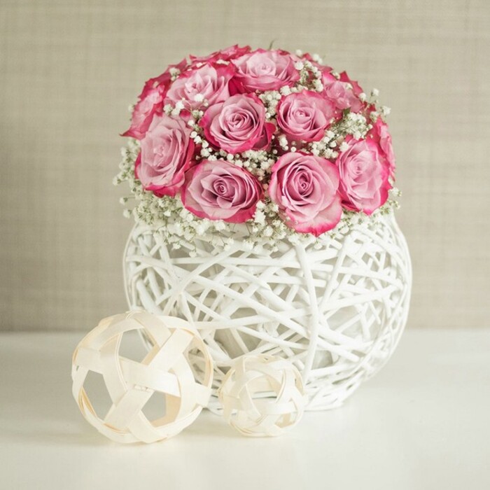 An artfully composed image centres on a generous bouquet of pink roses and white baby's breath arranged in a distinctive, spherical white vase, the sort of chic yet cosy design often chosen for homes around Queensbury and the surrounding streets. The bouquet forms a rounded, almost perfectly domed shape, with around fifteen to twenty roses clustered close together so that their petals overlap slightly. Each rose shows a subtle ombré effect: the innermost petals are a soft, powdery blush, gradually intensifying into richer, medium pink tones and finally into deeper, almost fuchsia edges along the outermost curls. Tiny clusters of Gypsophila are tucked between the roses, their fine white blooms creating a cloud-like halo that lightens the arrangement and gives it a romantic, celebratory tone, ideal for anniversaries, new baby greetings, or a quiet Valentine's surprise. The container is a globe-shaped vase crafted from white, interlaced strands reminiscent of rattan or wicker, forming an open, airy lattice. Light filters through these gaps, casting faint, criss-cross shadows onto the pristine white surface beneath, which could easily be a dining table or console in a Queensbury apartment. To the left of the vase sit two decorative spheres, one larger and one smaller, both spun from slender, natural-coloured wood or fibre strips twisted into an open, geometric pattern. Their warm beige notes complement the soft, neutral background: a gently textured wall in creamy beige that recedes quietly behind the scene. The lighting is bright yet diffused, without strong shadows, which flatters the roses' velvety texture and highlights the intricate weave of the vase and accents. The composition conveys tranquillity and modern elegance, suitable for illustrating home styling ideas, seasonal decor, or a refined floral gift chosen for same-day delivery to friends and family across Queensbury.