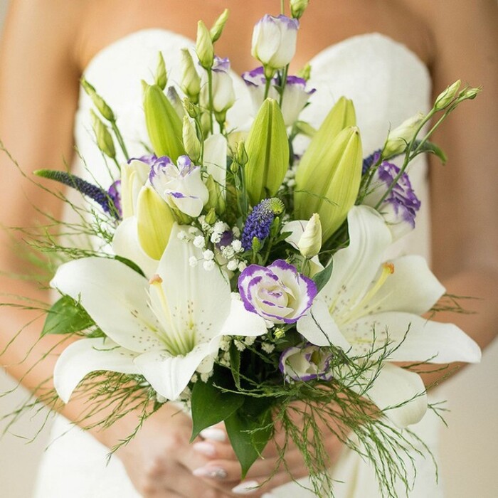 Captured in luminous detail, this Queensbury bridal bouquet reveals both artistry and a deep connection to local traditions. A cluster of fully opened white Oriental lilies anchors the design-their dramatic, curved petals feature delicate speckling and rich yellow stamens, each bloom exuding a sense of devotion against the bride's classic strapless gown. These lilies are joined by elegant white lisianthus, some fully bloomed with faint lavender frills, others in neat, promising buds. Intermingled throughout are slender, spiky clusters of indigo Veronica, their deep hues mirroring the vibrant purples found in Queensbury's springtime beds, and airy sprays of white baby's breath that lend the arrangement a soft, weightless quality. Broad, darker green lily leaves and swaying, pale green fennel-like foliage create a lavish, textured surround-each frond and leaf radiates freshness as if just gathered from a local flower market before dawn. The bride's light-polished nails gently enfold the sturdy stems, suggesting composure and reverence for the day ahead. A gentle blur of her white dress and bare shoulder behind the bouquet focuses all attention on this fusion of purity and vibrancy. Perfectly suited for brides seeking a blend of classic elegance and contemporary flair, this bouquet embodies the romantic, community spirit often celebrated at weddings in Queensbury's beloved venues.