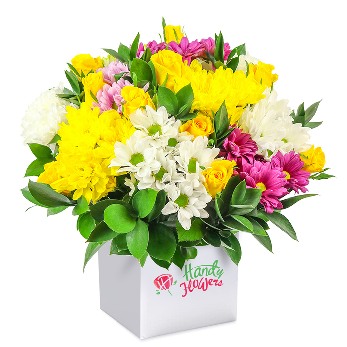 A generously filled, mixed flower arrangement is captured in a studio-style photograph, the bouquet overflowing from a pristine white square box and set against a completely white background that keeps all the focus on the blooms. The flowers are arranged into a smooth, dome-shaped canopy, giving the impression of abundance and careful craftsmanship. Dominating the colour palette are rich shades of yellow, from large, plush chrysanthemums with layers of fine, curling petals to clusters of compact yellow spray roses, some tightly closed and others just starting to reveal their centres. Their warm tones are cooled and balanced by groups of white chrysanthemums, including small, daisy-like blooms with bright green eyes and fuller, fluffier white varieties that add cloud-like texture. Woven through this cheerful mix are striking fuchsia and magenta chrysanthemums, some with lighter pink edges, which introduce depth and a modern burst of drama without overwhelming the softer colours. Smaller pink rosebuds and extra touches of yellow roses appear here and there, giving the composition a sense of movement and variety. All of the flowers emerge from a base of deep, glossy green foliage, whose sturdy leaves support the blooms and create a vivid contrast that accentuates the bouquet's brightness. The white cube container is minimalist and contemporary, its plain surfaces echoing the clean lines often favoured in Queensbury apartments and townhouses near the local parks. The mood of the arrangement is joyful yet refined, perfectly suited to sit on a dining table for a family celebration, to welcome someone to a new home in Queensbury, or to arrive as a surprise same-day delivery that instantly lifts the atmosphere of any room.
