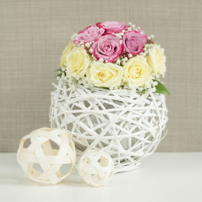 A beautifully styled floral centerpiece is shown in close detail, evoking the feel of a carefully prepared wedding or anniversary table in Queensbury. At the centre of the scene is a rounded bouquet of fresh roses, arranged so precisely that the blooms form a gentle, almost perfect sphere. About half of the roses are a soft pastel pink with the suggestion of lavender or fuchsia undertones, their petals smooth and velvety as they curl outward in layered spirals. The remaining roses are a creamy pale yellow, tipping into warm ivory, providing a gentle contrast that feels soothing rather than bold. Some blossoms are still partially closed, their centres tucked in like a secret, while others are more open, showing off the delicate architecture of each petal. Scattered among the roses are fine sprigs of white baby's breath, each tiny flower offering a crisp speck of brightness, filling the spaces between the larger blooms and creating a light, misty texture. The bouquet is set into a distinctive spherical vase made from white-painted wicker or delicate branches, woven into an open, cage-like pattern that allows a glimpse of the inner container and gives the piece a modern rustic charm. This style would suit many homes around Queensbury, from cosy terraces off Church Lane to airy new builds overlooking local green spaces. The vase rests on a bright white surface, perhaps a freshly laid tablecloth, its simplicity enhancing the sculptural quality of the arrangement. To the left lie two decorative spheres fashioned from pale, natural wood or rattan, one medium and one small, echoing the vase's weaving and creating a harmonious, curated display. A softly textured beige or light taupe backdrop, reminiscent of linen or subtle wallpaper, keeps the focus on the flowers while adding warmth. The overall impression is one of refined simplicity, a centrepiece perfect for thoughtfully designed gatherings or a calm everyday display in a Queensbury home.