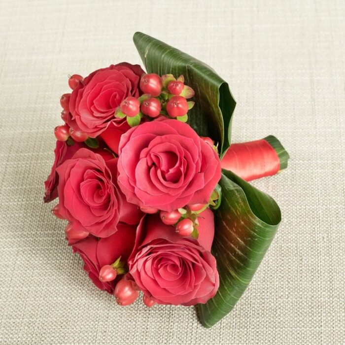 A lush cluster of deep red roses and berries has been arranged into a refined, compact bouquet and placed on a soft, light beige textile, whose subtle woven texture forms a gentle, unobtrusive backdrop. The bouquet itself features around six generously sized roses, each one fully open yet still holding a sense of structure, with velvety petals spiralling from tight, darker centers out to slightly lighter, gently curved outer edges. Between these rich crimson blooms, small bunches of round Hypericum berries in tones of red and reddish-pink are threaded throughout, adding tiny, polished spheres of colour that break up the expanse of petals and introduce a sense of movement and playfulness. Two or three broad, dark green tropical leaves sweep around the lower part of the arrangement, their glossy surfaces and clearly defined veins giving the piece a sculptural quality, as though the foliage is carefully cupping the flowers. At the base, the stems are drawn together and tightly wrapped with a deep red satin ribbon, the fabric smooth and subtly reflective, reinforcing the bouquet's polished, formal character. The lighting is calm and even, without harsh shadows, enhancing the feeling of intimate luxury and making the reds appear rich but not overpowering. This is the kind of design a Queensbury florist might create for a romantic gesture or as a bridesmaid bouquet for a local ceremony near the green spaces around Queensbury Circle: compact enough to hold comfortably, yet substantial and eye-catching. The overall mood is of timeless romance, ideal for anniversaries, engagements, or a same-day flower delivery that quietly says I love you without needing many words.
