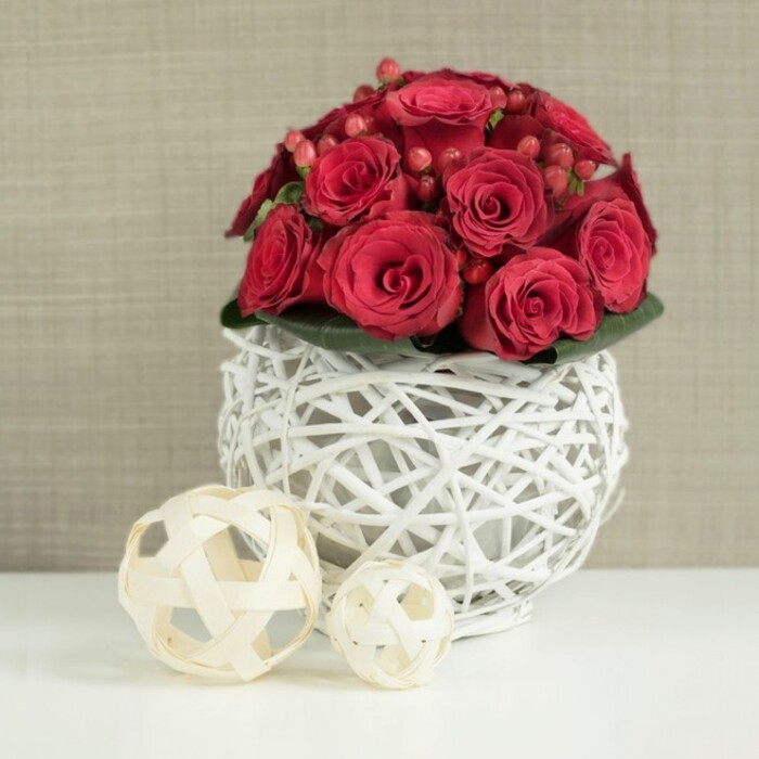 A beautifully composed wedding-style table arrangement of red roses fills the centre of the frame, set on a pristine white tabletop that enhances every nuance of colour. The roses are packed closely together in a neat, dome-shaped bouquet, their petals a deep, luxurious crimson that suggests passion and celebration. Some blooms are just beginning to unfold, their outer petals still curving protectively inward, while others are more open, revealing spirals of silky layers that catch the light. Among these roses, small clusters of red Hypericum berries add a different form of beauty: their smooth, rounded shapes and slightly brighter red tone create pockets of contrast and a hint of playful texture. Just beneath the flowers, a narrow ring of dark green foliage peeks out, offering a subtle natural border that gently emphasizes the fullness of the bouquet. The arrangement is nestled in a striking spherical vase made from white-painted twigs or wicker, woven into an open, basket-like globe. The structure feels both rustic and contemporary, with its pale lattice letting the white surface beneath show through. On the left side of the composition sit two decorative spheres made from light, natural-coloured wood strips, one slightly larger than the other, echoing the woven look of the vase but in soft beige tones. The background is a softly blurred wall in light taupe or beige, with a faintly textured or fabric-like finish that adds depth without drawing attention away from the flowers. The overall scene feels serene yet romantic, perfectly suited to a wedding reception at a Queensbury venue, an elegant proposal dinner at home, or any special occasion where a refined centrepiece is needed.