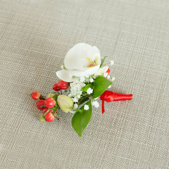 A meticulously arranged boutonniere is captured from above, resting on a light beige, woven linen-style fabric that provides a warm, neutral backdrop, similar to the simple table linens seen in local Queensbury venues. At the center of the composition is a flawless white Phalaenopsis orchid bloom, its broad, waxy petals gently spreading outwards in a smooth oval, subtly curved at the edges. Within the orchid's throat, tones of creamy yellow and soft orange emerge, giving the flower a delicate heart of colour that suggests both warmth and refinement. Surrounding this central orchid, airy tufts of white baby's breath form a halo, their tiny clustered florets creating a cloud-like softness and an almost misty effect around the more defined petals. To one side, a tight cluster of glossy, bright red berries catches the eye, each small sphere reflecting light and introducing a bold, festive contrast that would feel perfectly suited to a winter wedding or seasonal party near Queensbury Park. A few pale green, oval buds are nestled among them, adding freshness and a sense of living growth. Interlaced throughout the design, lush green foliage in varied silhouettes-some leaves broad and pointed, others slender and elongated-frames the whole piece with a rich, natural depth. The stems are carefully bound at the base with a rich red satin ribbon, wrapped snugly and tied in a neat, understated knot, its short tails lying flat against the fabric. This polished finish makes the boutonniere appear ready to be pinned to a groom's jacket or a prom suit for a special event in Queensbury, combining classic white, green, and red in a small but exquisitely detailed floral accent.
