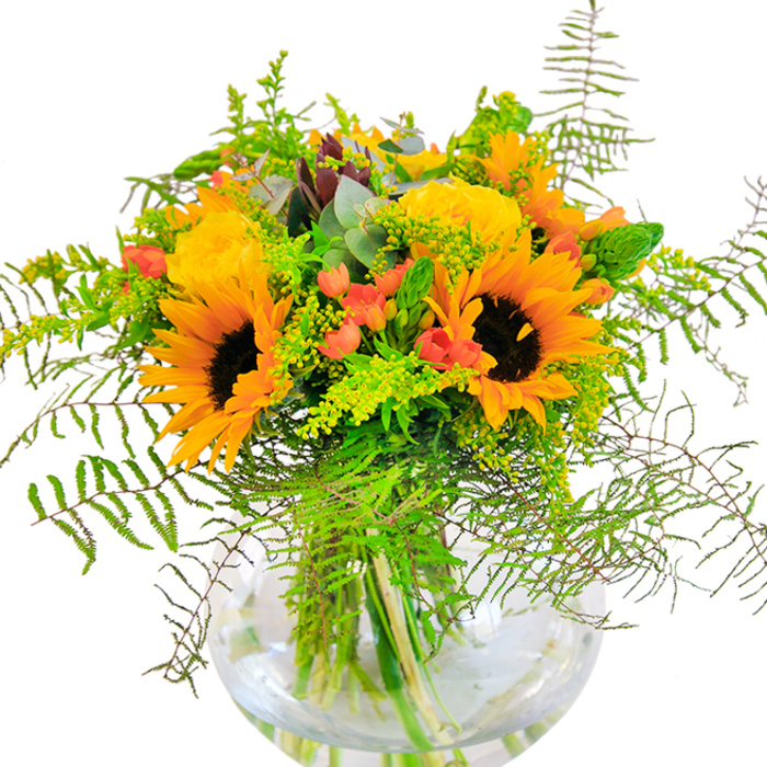 Radiating a spirit of joyous abundance, this striking arrangement weaves together the vivid beauty of late-summer florals with the familiar sense of community found throughout Queensbury. A constellation of bright sunflowers provides an exuberant focal point, their golden-yellow petals and striking dark centers conjuring images of sun-drenched gardens beside the Jubilee line. Nestled among them, rich yellow garden roses reveal their intricate spiral formations, offering plush texture and a classic sophistication. Glossy, scarlet hypericum berries add delightful pops of colour, reminiscent of berry-picking excursions in local parks. The arrangement is embroidered with layers of greenery-airy ferns cascading downwards, slender green spikes shooting upwards, and sprigs of delicate goldenrod dotted with tiny yellow blooms. Wisps of silvery eucalyptus leaves add a soft, aromatic touch, all gathered in a transparent, round glass vase where stems are elegantly displayed in clear, fresh water. The interplay of colour and texture suggests a celebration worthy of milestone birthdays, anniversaries, or festive gatherings in Queensbury households. The bouquet's visual vibrance is matched by an undercurrent of gentle, uplifting fragrance, and the tactile mix of soft petals and crisp foliage. Meticulously crafted with local knowledge and artistry, this arrangement channels the heartening, sunlit essence of Queensbury, making it a meaningful gift or a stunning centrepiece for any home.