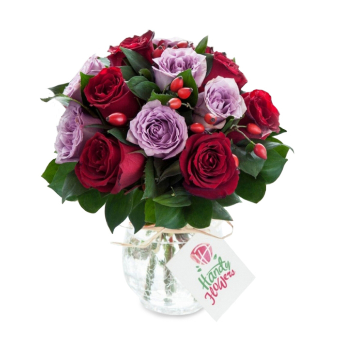 A densely gathered, romantic bouquet of roses in rich, contrasting tones-deep crimson and burgundy paired almost equally with soft lavender and dusty purple blooms-designed to read as both elegant and warmly intimate. Each rose is fresh and fully open, petals layered in delicate spirals that suggest velvet under the fingertips; the darker roses bring a sense of drama while the lavender varieties add a soothing, soft edge. Interspersed throughout the cluster are glossy dark-green leaves, likely Ruscus, which lift and separate the blooms so they read clearly as individuals while still forming a cohesive round shape. Small, bright red-orange rose hips are dotted through the bouquet, offering a rustic, tactile counterpoint that adds movement and seasonal charm. The stems are held in a rounded, bulbous glass vase with a mercury or crackled silver finish that catches light and reflects subtle glints across the petals; a length of natural twine is tied at the neck and a small rectangular white gift tag hangs forward with a hand-drawn floral motif and handwritten space for a message. The arrangement is set against a pristine white background that makes every hue sing and emphasizes the professional finish. Prepared with neighbourhood care by Flower Delivery Queensbury, this bouquet suits anniversaries, congratulatory gestures, or those moments when a luxurious, fragrant floral gift is needed across Queensbury, Kingsbury and the approach to Wembley - a composition that feels both refined and warmly local.