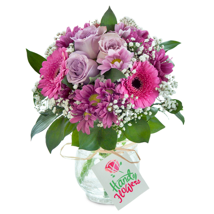 Centered in the soft, defused light of a studio setting, this exuberant bouquet exemplifies the vibrant floral artistry found in Queensbury. Soft lavender roses are gathered in tight, elegant clusters at the heart of the arrangement, each bloom shimmering with freshness as if plucked at dawn. Encircling the roses, striking hot pink Gerbera daisies beam with energy-their bold petals fanning out from deep, dark centers. Alongside them, generous sprays of deep magenta chrysanthemums, their petals tipped with a slight greenish-yellow, contribute visual depth and a rich, plush texture. Wisps of dainty white baby's breath drift between the main blossoms, lightening the composition and evoking a fresh spring breeze, reminiscent of walks through Gower Road's lush greenways. Glossy dark green leaves nestle at the base, providing contrast and a sense of fullness. The entire bouquet is presented in a rounded, clear glass vase with a cracked glass texture that sparkles subtly, reflecting light in a way that's both rustic and refined. A crisp raffia ribbon is tied neatly at the vase's neck, completed by a rectangular white tag thoughtfully designed for a message or dedication. Each element is carefully chosen to create a harmonious palette of purples, pinks, and greens, ideal for gifting on anniversaries, birthdays, or brightening a home in Queensbury's lively community. The arrangement reflects the dedication and flair of local floral designers, making it a perfect embodiment of special occasion gifting and everyday beauty.