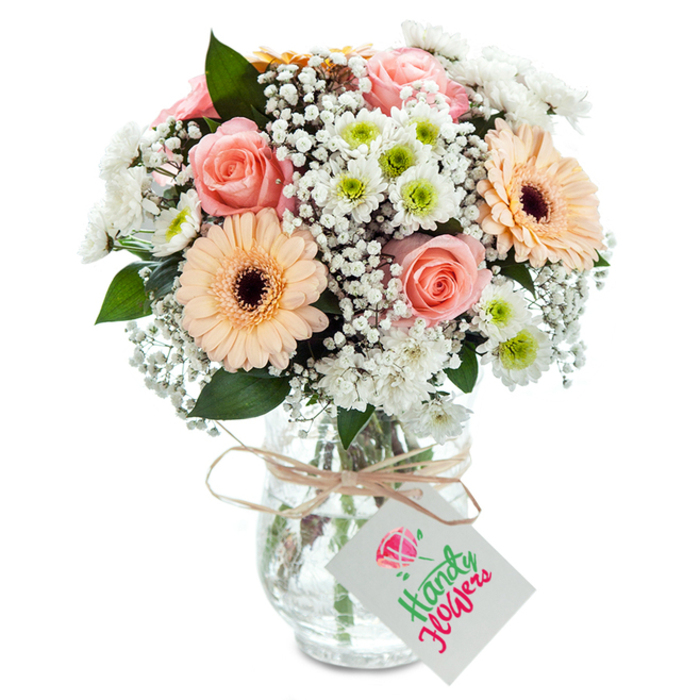 Graceful and inviting, this local bouquet captures the easygoing spirit of Queensbury with its artful blend of textures, hues, and thoughtful touches. At the heart, plump, pastel-hued roses unfurl with a classic elegance, casting warm peach-pink tones through the arrangement and evoking strolls along Queensbury's leafy avenues. Flanking these are a pair of vivid peach gerberas, their broad petals and deep brown centres providing cheerful counterpoints and a sense of celebratory flair. Sprigs of white baby's breath float between, creating a light, cloudlike background that breathes air into the composition. Crisp white chrysanthemums with lively green centres echo the freshness of neighbourhood gardens, while hints of eucalyptus foliage add a layer of verdant scent and natural texture. The bouquet sits gracefully in a clear glass vase with gentle texture, stems arranged in neat, visible clusters and the water sparkling below. A rustic twine bow and a blank, crisp tag dangle at the neck, inviting your personal message-an understated, elegant detail. With its mastery of colour and balance, this arrangement is the perfect floral gift for any occasion across Queensbury, from birthdays to get-well wishes, or simply offering a bouquet of everyday beauty.