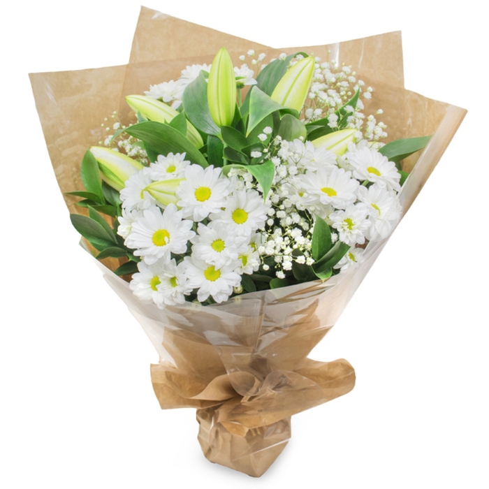 A carefully composed hand-tied bouquet sits upright against a clean white field, the arrangement's white and green palette presented with restrained elegance that would read well on a mantel in a Queensbury home. Dominating the composition are numerous white, daisy-like chrysanthemums: small, dense heads of many symmetrical petals surrounding subtle yellow centres, which give the bouquet a classic, uplifting presence. Threaded through these are several elongated lily buds in a pale greenish-yellow tone, a couple with faint striping along their flanks; a few of the lilies are just beginning to part at the tips, imparting a sense of quiet promise. Fine sprays of gypsophila punctuate the spaces between larger blooms, their minute white flowers forming a soft, cloud-like veil that adds delicate texture without overwhelming the main shapes. Deep green foliage-broad leaves and glossy stems-frames and grounds the blooms, emphasizing the clarity of the whites. The flowers are gathered and wrapped in two distinct layers: an inner sheet of rustic, crinkled brown kraft paper that lends an organic, market-fresh feel, and an outer layer of clear cellophane that catches the light and protects the arrangement during handling. The base is neatly secured, forming a compact, portable bouquet that is both practical and refined. This composition conveys serenity, purity, and thoughtful care, suitable for a range of moments from birthdays and thank-you gestures to sympathetic remembrances across Queensbury, Harrow and neighbouring Wembley.