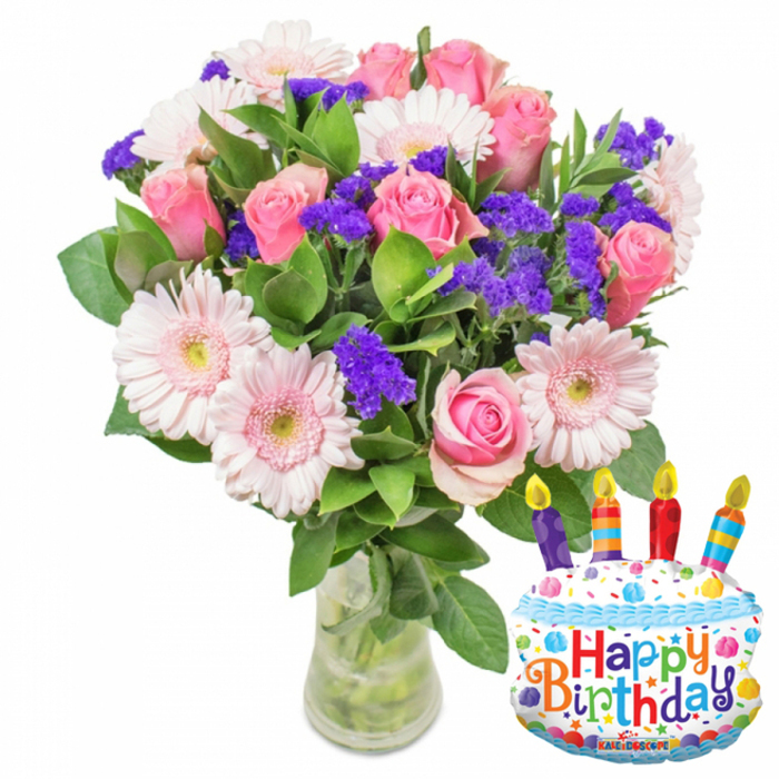 A bright, carefully composed birthday arrangement occupies the left side of this image, arranged in a simple clear glass vase and set against a crisp white background that keeps every colour true. The bouquet is dominated by fully bloomed soft pink roses, their velvety petals layered and slightly open, partnered with pale pink Gerbera daisies whose broad petals form sunny circles with pale yellow-green centres. Nestled between the larger flowers are abundant sprays of vivid purple statice or limonium, their tiny clustered blooms adding contrast and a sculptural texture that lifts the composition. Small green-leaved stems and glossy foliage weave through the stems, giving weight and structure while the clear vase shows the organised tangle of green stems and fresh water beneath. To the right a metallic foil balloon shaped like a layered birthday cake provides a festive counterpoint: white frosting-like panels, blue piping along the edges and a joyful dusting of red, yellow, green and blue sprinkles; four multi-coloured striped candles in shades of red, orange, blue and purple stand at the top with painted yellow flames, and the message 'Happy Birthday' is printed across the front in bright, playful letters. A border of tiny stars and confetti patterns decorates the balloon's bottom edge and a small 'KALEIDOSCOPE' logo is visible along the base. The arrangement radiates a feeling of thoughtful celebration - suitable for a birthday surprise delivered locally to homes around Queensbury, Wembley Park or neighbouring Kingsbury.