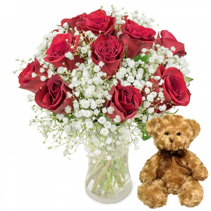 This carefully lit composition presents a classic romantic gift set, captured as though ready to leave a Queensbury florist's bench for same-day delivery. Dominating the left side of the frame is a luxurious bouquet of approximately twelve deep crimson red roses, gathered in a simple clear glass vase. Each rose is lush and full, with velvety petals that spiral outward in neat layers, some blooms perfectly open and others just beginning to unfurl, adding natural variation and depth. Threaded between these passionate red blossoms are generous clusters of white baby's breath, their countless tiny flowers creating a soft, misty surround that lightens the overall look and gives the bouquet an airy, cloud-like volume. The contrast of the dark red petals, snow-white gypsophila, and glimpses of dark green foliage evokes the type of refined designs often chosen in Queensbury for anniversaries, engagements, and milestone birthdays. Through the vase you can see the straight green stems immersed in clear water, emphasising freshness and professional arranging. To the right of the flowers, a charming teddy bear in light brown plush sits facing forward, positioned as if waiting on a table in a home off Streatfield Road. The bear's fur appears wonderfully soft and slightly tousled, its round black button eyes and small black nose giving it an endearing, almost childlike expression. A neatly tied dark brown satin ribbon encircles its neck, adding a simple but elegant touch. With its serene white background, the image highlights the emotional warmth of this pairing of roses and teddy bear, making it an ideal visual representation of romantic gestures and thoughtful gift delivery around Queensbury.
