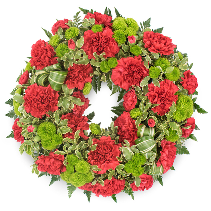 The image presents a perfectly round sympathy wreath laid out against a stark white background, the clean setting drawing full attention to its lush ring of red and green. The wreath is tightly packed with deep red carnations, each bloom displaying intricately rippled petals that overlap like layers of delicate fabric, creating a feeling of depth and softness all around the circle. By contrast, vivid lime green button chrysanthemums are dotted throughout, their small, spherical flower heads neatly formed and almost moss-like in texture, adding energetic flashes of colour that lift the design. The flowers sit on a structured bed of greenery that both supports and frames them: dark leatherleaf-style fern fronds radiate outward, their fine leaflets forming a feathery outline, while glossy variegated pittosporum leaves with creamy margins weave through the flowers, breaking up the solid colours and offering a fresh, garden-like look. Tucked into the lower right side is a decorative bow created from a wide, striped green ribbon, the loops curling gently over the foliage and echoing the lime tones of the chrysanthemums. The craftsmanship feels deliberate and careful, as if arranged by a local Queensbury florist used to creating meaningful tributes for services near community churches and crematoriums around the area. The lighting is bright and neutral, leaving the colours true and the textures sharply defined-every ruffle of carnation, every rounded button mum, and the subtle sheen of the ribbon are clearly visible. The overall mood is one of respectful remembrance, with the vibrant reds symbolising love and the greens hinting at renewal, making it an appropriate funeral or memorial wreath for families across Queensbury who wish to send a heartfelt, traditional floral farewell.