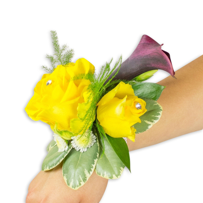 Seen close-up on a light-skinned forearm against a pure white studio background, this hand-tied wrist corsage balances brightness and drama with confident craftsmanship. Two vivid yellow roses take centre stage-the left bloom wide and open, the right a little more buttoned-each softened with layered petals and highlighted by a small clear rhinestone nestled toward the center, adding a pinpoint of celebratory sparkle. Behind and slightly above the roses a deep burgundy to plum calla lily arcs gracefully, its smooth, satiny surface and trumpet-like form creating a sophisticated counterpoint to the round, joyful roses. The composition is grounded by a collar of foliage: broad variegated leaves with pale margins (reminiscent of pittosporum) provide structural weight while darker green leaves anchor the lower edge; airy asparagus fern threads through the top to give lift and delicate texture. Tiny white filler flowers nestle among the foliage, filling gaps and lending an overall soft, garden-made feel. The corsage is worn diagonally across the wrist and secured with a slim ribbon or elastic that remains mostly concealed beneath the blooms. As a local Queensbury florist I picture pairing this compact design with suits and gowns for prom or wedding parties, or arranging similar pieces for clients collecting from our shop near Queensbury station and Fryent Country Park. The combination of sunny yellow, deep plum, and fresh green reads bright, elegant, and unmistakably celebratory-ideal for marking momentous family occasions throughout Queensbury and nearby Wembley.
