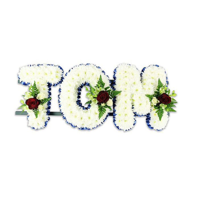 A custom sympathy tribute created by Flower Delivery Queensbury presents the name TOM in large, three-dimensional letters densely covered with tiny, pristine white chrysanthemums. The chrysanthemums are arranged for maximum pillowy volume, each petal layer contributing to a seamless, soft surface that captures cool studio light. Each letter is edged with a striking royal blue ribbon that crisply defines the silhouette and adds a formal, comforting accent against the white blooms. Within the heart of every letter a carefully composed floral spray provides a deliberate point of focus: a single, deep dark red rose in each cluster symbolizes love and remembrance, while smaller creamy-white spray roses and slender fern fronds soften the composition and introduce gentle green undertones. The foliage is chosen for texture and longevity, its matte leaves contrasting with the delicate shine of rose petals. A thin, dark metal bar at the base and slight framework behind the letters gives subtle support, intended for upright placement at a funeral venue or remembrance display, and is visible only as a low line beneath the arrangement. Set against a clean white background, the design reads with calm dignity-colours and shapes clear and readable for mourners across the room. This handcrafted name tribute is suitable for services in Queensbury and nearby Kingsbury or Wembley, and it reflects a local florist's understanding of ceremony: respectful, carefully balanced and made to comfort those gathered in farewell.