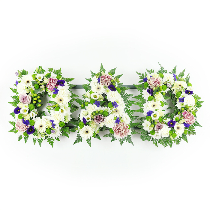 A poignant floral display forms the word DAD in three solid, capital letters, laid out horizontally and photographed against a bright, uncluttered white background that makes each colour and texture shine. Each letter is created from a dense bed of pristine white chrysanthemums and similar daisy-headed blooms, so closely arranged that they resemble a soft, continuous blanket of petals. These white flowers provide a serene foundation, evoking purity, peace, and the quiet stillness that often follows a loss. Threaded through this snowy base are soft accents of colour: gently opening roses in shades of lavender and pale mauve, ruffled lilac carnations, and perhaps the occasional bud just beginning to unfurl. Deeper purple notes, suggestive of lisianthus or pansy-like blossoms, are positioned to catch the eye without overwhelming the calmness of the whites, adding a regal, respectful tone. Around and between the letters, a Queensbury-based florist has framed the design with lush greenery-feathery fern fronds, rich green leaves, and small clusters of bright green button chrysanthemums that shape and support the letters. This foliage traces the curves of the letter D and the angles of the A, providing structure and a natural contrast, much like the patches of green that break up the urban streets around Queensbury station. The lighting is soft yet clear, emphasising the careful craftsmanship, from each flower's placement to the neat, even outline of the tribute. It is an eloquent, personalised funeral arrangement, perfectly suited for a memorial or crematorium service in the Queensbury area, honouring a beloved father with grace and heartfelt sincerity.