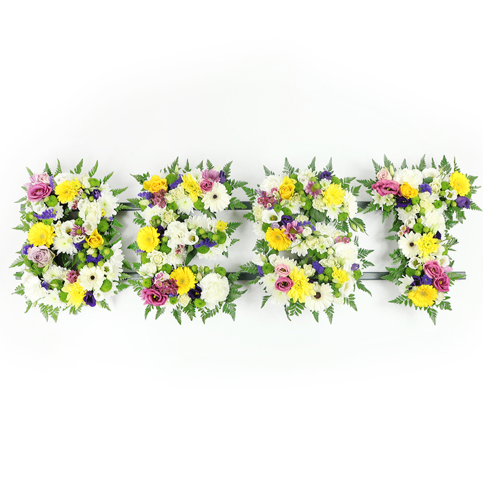 This head-on photograph shows the word BEST rendered in four solid, flower-covered letters, each a compact tapestry of colour and texture set against a bright white studio background. The overall palette leans toward cheerful yellow and soothing white: sunny yellow roses and gerbera provide luminous focal points, while fluffy white chrysanthemums and cream-toned gerberas soften the composition and evoke purity. Interspersed throughout are soft purple lisianthus or similar petite blooms, and pops of vivid pink and magenta from spray roses or carnations add a touch of celebratory warmth. Small clusters of pale green and white filler blooms nestle between the main flowers, giving the surface a tactile, layered quality that invites touch. Lush dark-green fern fronds flank each letter, their feathered edges creating a gentle, botanical border that contrasts elegantly with the bright petals. In places a narrow dark-green or metal support bar is visible, hinting at the careful armature supporting this large-scale tribute. The lighting is even and clear, designed to showcase the fresh textures and the subtle sheen of petals and leaves, while a faint floral scent might be expected in person. As a local florist working around Queensbury, I can picture this arrangement positioned at a memorial service on the town High Street or delivered to a family home after a service nearby, where its uplifting colour and considered craftsmanship will convey love, respect and remembrance in equal measure.