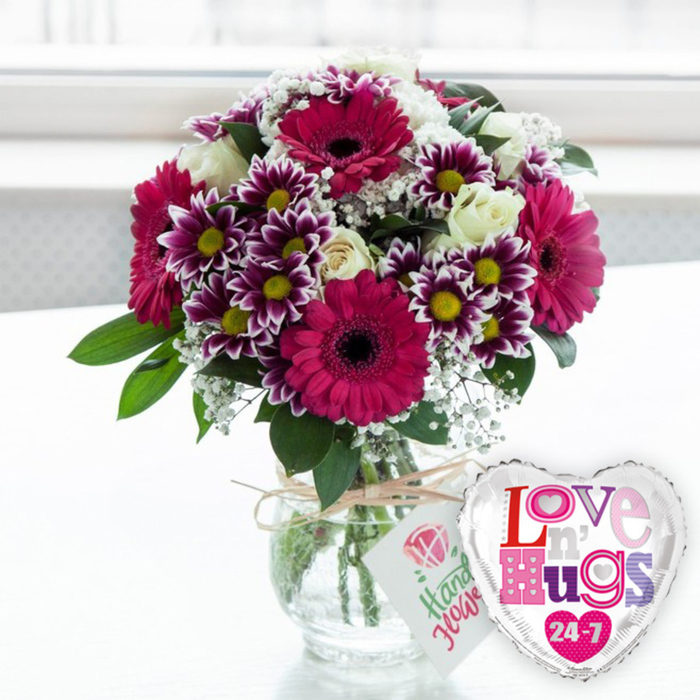 A joyful floral display in a clear glass vase is paired with a playful heart-shaped balloon, creating an image that feels like a surprise delivery to a cosy Queensbury flat. The bouquet itself is rich and abundant, built around several large gerbera daisies in striking fuchsia and magenta tones; their bold, dark centres and layered petals give a modern, graphic feel. Intermixed with these are numerous chrysanthemums whose petals shift from deep, royal purple at the base to crisp white at the tips, the golden-yellow centres shining like tiny suns and adding a spark of cheer. Between the more vivid blooms, creamy white roses rest in varying stages of opening, their velvety petals gently spiralling inward, bringing a sense of timeless elegance. Airy sprays of white baby's breath thread through the arrangement, soft feathers of blossom that create lightness and cushion the stronger colours, while glossy green leaves peek through like glimpses of a spring garden. The stems of all the flowers are clearly visible through the rounded, transparent vase, cut at an angle and arranged carefully in clear water that hints at freshness. A length of rustic natural twine is wrapped around the neck of the vase and tied neatly, holding a small rectangular white tag that bears a pink rose illustration and casual green script suggesting a handmade, personal touch often associated with local florists near Honeypot Lane and the centre of Queensbury. To the right of the vase, slightly overlapping the base, is a gleaming silver foil balloon in the shape of a heart, its reflective edge framing a white panel where colourful, patterned letters spell Love n' Hugs 24-7, scattered with small hearts. The entire scene rests on a clean, pale surface, while the background dissolves into a soft blur of white light, evoking a bright window and giving the arrangement an uplifting, celebratory atmosphere perfect for birthdays, anniversaries, or a spontaneous show of love.