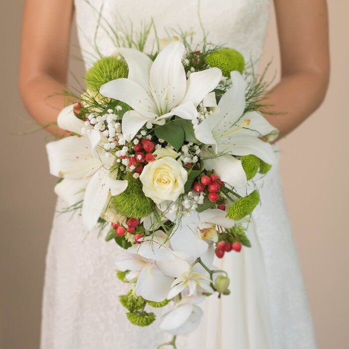Captured in luminous detail, this bridal bouquet held against the creamy lace of an ivory wedding gown is a celebration of texture, movement, and colour, thoughtfully crafted by a florist familiar with the spirit of Queensbury. Large, trumpet-shaped white lilies dominate the design, their petals brushed with understated speckles, radiating a sense of refined opulence. A single, rich cream rose sits at the centre, tightly swirled to convey a sense of anticipation and enduring love. Bright lime-green pom-pom chrysanthemums are tucked artfully among the white blooms, their vivid shade offering a burst of life akin to the lively market days along Queensbury Circle. Throughout the bouquet, soft baby's breath dances between stems, while clusters of lustrous red Hypericum berries provide a playful nod to local winter celebrations. The composition's dramatic teardrop silhouette is crafted by cascading Phalaenopsis orchids, whose pure white blooms add an airy, modern edge, gently trailing past the bride's hands. Whisps of grass and lush deep green foliage anchor the bouquet, harmonising nature's loose artistry with precise craftsmanship. The soft, out-of-focus backdrop draws attention to each floral nuance, highlighting this lavish display as a quintessential choice for spring or summer ceremonies in Queensbury or for brides desiring a fusion of classic romance and fresh, seasonal flair.