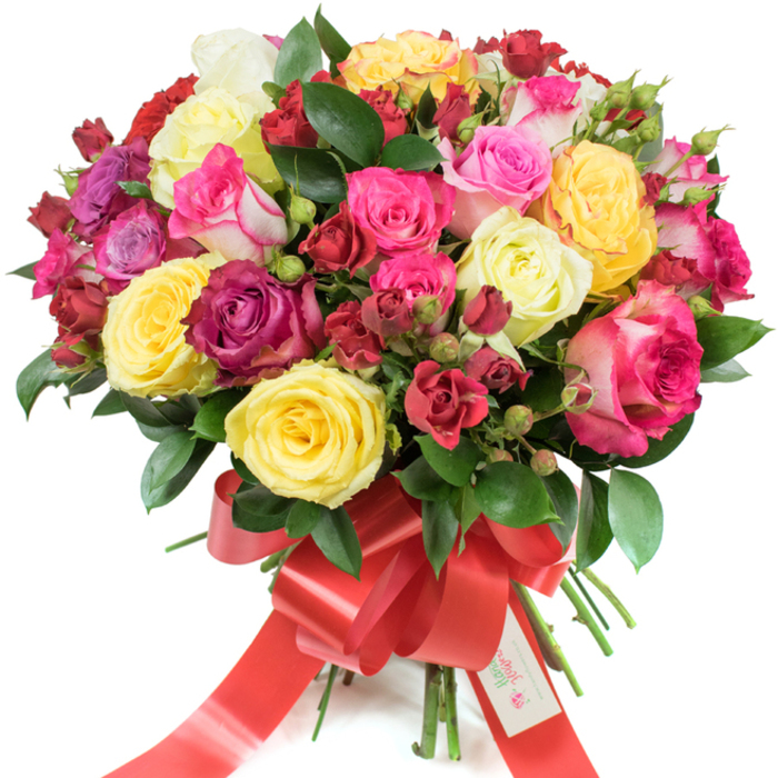 A richly coloured, professionally arranged bouquet of roses is shown against a stark white background, its rounded profile full and deliberately dense to showcase the variety of tones and textures: bright yellow roses that catch the eye, creamy ivory blossoms, a spectrum of pinks from soft blush to intense fuchsia and delicate pink-and-white bi-colours, balanced by deep burgundy and crimson clusters. The mix includes fully open hybrid tea roses with layered, silky petals, smaller spray roses that form compact clusters, and fresh tight buds that promise continued bloom. Interspersed throughout is glossy, dark green foliage - likely ruscus or salal - which provides structure and a cooling visual counterpoint to the warm floral hues. The stems are neatly bound and finished with a wide red satin ribbon tied into an elegant bow whose long tails cascade down; a small white tag bearing a logo and the words Happy Holidays is secured at the knot, suggesting a seasonal or celebratory intent. The lighting is gentle and even, allowing the satiny surfaces and subtle veining of petals to become visible; one can almost detect a faint citrus-tinged rose perfume in the air. As crafted by a florist familiar with Queensbury and nearby Kingsbury, this bouquet reads as an opulent, joyful gift suitable for special occasions - a statement of love, congratulations, or festive cheer delivered with local care.