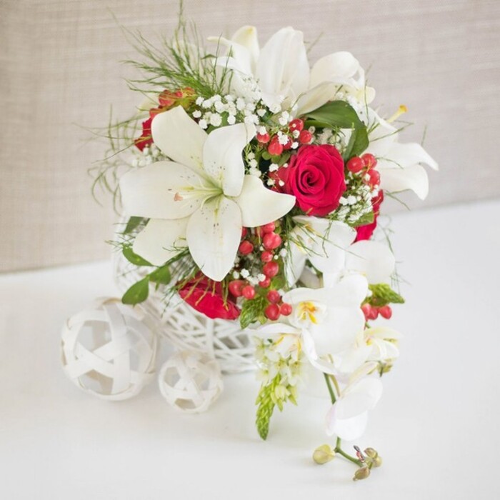 A sophisticated floral display is styled as a wedding or special-occasion table arrangement, poised on a smooth white surface against a softly textured beige backdrop reminiscent of a calm, sunlit wall in a Queensbury home. At the heart of the scene is a round, white basket-style container with an intricate open lattice, its airy weave suggesting hand-crafted detail that feels right at home near the boutiques and cafés around Queensbury Station. From this sculptural base, large white oriental lilies rise and tilt at different angles, their broad, six-petalled blooms open wide, with golden stamens and gentle shadows pooling in the throats of the flowers. Between them, velvety red roses offer a deep, romantic counterpoint, their petals layered and lush, catching the light with a subtle sheen. Delicate strands of white baby's breath float among the larger blooms like small clouds, softening the transitions between white and red. Fine green foliage, with a feathery, almost airy quality, threads through the design, adding texture and a garden-fresh feeling akin to walking through one of the green spaces near Kingsbury. Here and there, clusters of glossy red hypericum berries tuck into the arrangement, their rounded forms lending a festive accent that makes the piece suitable for winter weddings or holiday celebrations in and around Queensbury. Cascading elegantly from one side, a line of white Phalaenopsis orchids spills downward, each orchid bloom with a nuanced touch of yellow and pink at the centre and a few buds held in reserve. On the left, several white woven decorative spheres rest on the table, mirroring the basket's texture and visually balancing the orchids' gentle descent. The composition exudes romance, refinement, and celebration, ideal as a focal point for a wedding table, anniversary dinner, or stylish event in the local area.