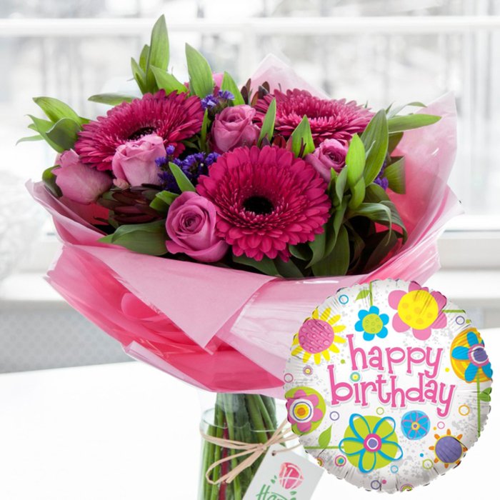 A beautifully styled birthday flowers and balloon display is shown in soft, natural indoor light, as if set on a table in a Queensbury home near the open spaces of Eton Grove Park. At the heart of the composition stands a clear glass cylindrical vase, its smooth surface reflecting the light and revealing the neatly trimmed stems inside. Rising from it is a generous bouquet dominated by vibrant pink tones: large Gerbera daisies in rich fuchsia and magenta stretch out wide, their petals forming sunburst discs around dark, almost black centres that add drama and depth. Clustered among them, numerous pink roses in varying stages of bloom lend an air of gentle romance, from tight, rounded buds to partially opened flowers showing soft, layered petals with a delicate, velvety texture. Sprigs of small, deep purple filler flowers - likely statice - weave through the arrangement, adding fine detail and a cool contrast to the warmth of the pinks. The bouquet is framed by abundant green foliage, broad and narrow leaves mingling together to create fullness and a lush, garden-fresh feel, as though prepared moments ago by a local Queensbury florist. At the base of the display, translucent pale pink cellophane or tissue is gathered and wrapped around the stems, spilling slightly over the vase rim to create a soft, cloud-like collar. A simple off-white rectangular tag, printed with a subtle rose emblem, dangles from rustic twine tied neatly around the vase neck, suggesting a thoughtful, high-quality birthday gift. In front, slightly to the right, a round foil Happy Birthday balloon floats, its white and lightly speckled surface adorned with playful, colourful floral and circular motifs in yellow, sky blue, lime green, vivid pink, and deep purple. Bold pink script across the centre reads happy birthday, echoing the tones of the flowers. The softly blurred background suggests a bright, homely Queensbury interior, keeping all attention on this joyful, ready-to-deliver birthday surprise.
