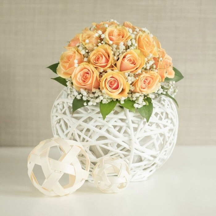 A refined floral centerpiece of peach-toned roses arranged in a perfect dome rests on a bright white surface, as though prepared for an elegant gathering in a Queensbury dining room. Approximately two dozen roses are packed closely together, their heads forming a rounded mound of colour that shifts from deeper apricot and soft tangerine at the centres to paler, almost buff-cream at the outer petals. Each rose is at a slightly different stage of opening, some still cupped and promising, others more open and layered, which gives the bouquet a natural, living character. Between the roses, airy sprays of white baby's breath weave in and out, creating a delicate mist that softens the overall look and adds a gentle, romantic texture. Tucked beneath are glossy, dark green leaves that frame the base of the blooms and add depth, reminiscent of fresh foliage you might see in gardens along local Queensbury streets. The arrangement is held in a striking, round vase constructed from white woven strips, forming a criss-cross lattice that looks simultaneously sculptural and light. This open, basket-like structure allows glimpses through to the interior and creates interesting shadows where light filters through. In the foreground, resting on the same white tabletop, two smaller decorative spheres echo the woven motif. One is slightly larger and a warm beige tone, the other smaller and cream, both created from slender, interlaced strips in an open geometric pattern. Behind the display, a softly textured, pale beige wall provides a calm, uncluttered background that enhances the sense of quiet sophistication. The gentle, diffused lighting emphasizes each petal and woven line, suggesting a setting ideal for wedding décor, home staging, or a thoughtful floral delivery for a special moment in Queensbury.