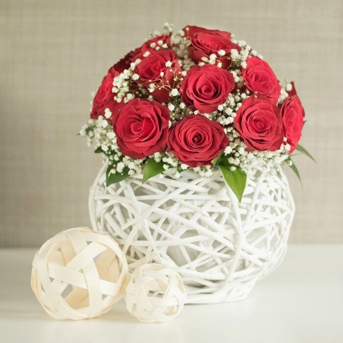 A sophisticated floral centrepiece featuring deep red roses and delicate white baby's breath is displayed on a pristine white surface, evoking the polished calm of a carefully dressed wedding table in Queensbury. The flowers form a compact, spherical bouquet of roughly twelve to fifteen roses, each bloom open and velvety, with petals tightly layered in elegant spirals. Their rich crimson tone offers a sense of classic romance, ideal for a bridal table or an anniversary meal in a home near Queensbury Circle. Nestled amongst the roses, fine stems of baby's breath scatter clusters of tiny white blossoms, creating a soft, cloud-like halo that contrasts beautifully with the bold red. A few subtle green leaves peep through the flowers, adding just enough foliage to remind you that these are fresh stems, prepared by an experienced local florist who understands understated luxury. The bouquet is cradled in a striking round white vase with an open, woven structure, its slender strands intertwining to form a light, airy lattice reminiscent of handcrafted wicker decor found in stylish townhouses around Honeypot Lane and Edgware. Resting on the same white tabletop to the left of the vase are two decorative spheres made from pale, flat woven strips, one noticeably larger and placed closer to the viewer, the smaller one positioned just behind. Their openwork design harmonises with the vase and contributes to a cohesive, contemporary look. Behind the arrangement, a softly textured, neutral-toned wall fades gently out of focus, framing the piece without distraction. The overall impression is one of tranquil elegance and romantic warmth, perfectly suited to wedding table arrangements, engagement dinners, or refined home styling across the Queensbury area.