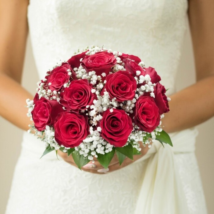In the soft, inviting light of a Queensbury morning, a bride pauses, bouquet held lovingly at her waist, offering a perfect snapshot of wedding-day grace and floral artistry. The bouquet itself commands attention-a domed masterpiece featuring dozens of rich, red roses, each petal closely layered to create a tapestry of velvety color and romantic depth. Sprinkled throughout, clusters of white baby's breath float like small clouds, their airy delicacy setting off the intensity of the roses and echoing the bouquet's timeless allure. A few stems of vibrant green foliage peek from underneath, suggesting freshness and natural elegance inspired by Queensbury's many green spaces and community gardens. The bride's hands, adorned with manicured nails, curve gently around the bouquet's stem, a tender gesture that speaks of anticipation and love. Behind the blooms, the soft curves and intricate textures of an ivory or white wedding gown are partially visible-the bodice possibly a swirl of lace or embroidered detail, and a whisper of softly draping fabric trailing along the side. The background is rendered in gentle, creamy tones, causing the vivid colours and fine textures to pop in clear relief. This composition radiates a warm, celebratory mood, making it an ideal visual for couples dreaming of their wedding in Queensbury or for articles highlighting local wedding flowers and traditional bridal bouquets signed by the skillful hands at Flower Delivery Queensbury.