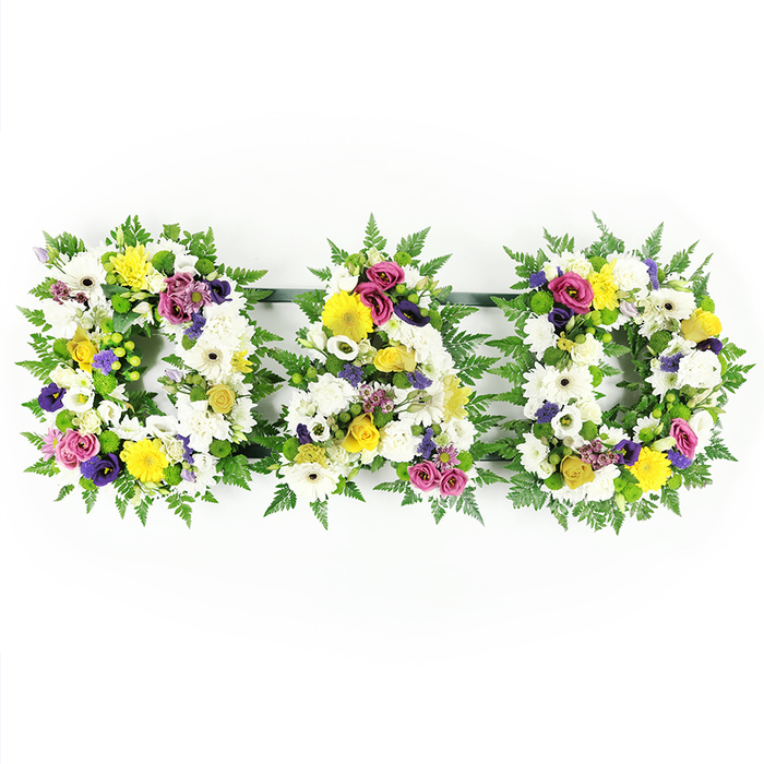 In this image, a traditional funeral flower tribute spells out the word DAD in three bold capital letters, designed with the quiet elegance that many families around Queensbury look for when honouring a father's memory. The letters - D, A, and D - stand in a neat row on hidden dark green supports, giving the impression that they are floating together as one unified piece against the bright white background. Each letter is formed from a base of rich, verdant fern foliage, the fronds overlapping and curling around the edges to create a deep green border that frames the flowers and adds a natural softness. Filling the centre of every letter is a dense carpet of white blooms: classic chrysanthemum heads and large, open gerbera daisies, their crisp white petals layered tightly so that no gaps interrupt the solid shape of the word. Among these, the florist has tucked clusters of sunny yellow gerbera daisies and delicate yellow spray roses, adding warmth and hope to the otherwise serene palette. To enrich the design, accents of purple lisianthus, deep magenta or fuchsia-toned spray roses, and tiny lavender flowers are placed thoughtfully around the letters, while bright green button chrysanthemums offer small pops of fresh colour and texture. The composition is front-facing, symmetrical, and carefully balanced, ideal for display at a funeral, memorial, or graveside service near Kingsbury, Queensbury Circle, or along Honeypot Lane. The overall mood is one of respectful remembrance, tenderness, and gratitude, conveying the simple yet powerful message that a much-loved dad is being honoured with care and artistry through fresh flowers.