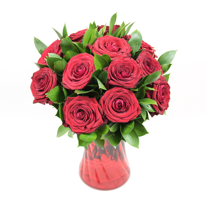 In this image, a beautifully composed bouquet of about a dozen deep red roses is arranged in a perfectly rounded cluster and displayed in a translucent red glass vase, all set against a stark white background that makes the colours glow. Each rose is fully open yet still crisp, with velvety petals layered so tightly they form intricate spirals, drawing the eye into the centre of every bloom. The red is a rich, saturated crimson, with subtle variations in tone that give the flowers depth and a sense of three-dimensional fullness, as though you could almost feel the softness of the petals under your fingertips. Nestled among the roses is a generous amount of dark green foliage, likely ruscus or a similar leaf, with smooth, lance-shaped leaves that shine with a gentle gloss. These greens peek out at different angles, adding texture, movement, and an organic frame around the rose heads, as if carefully placed by a skilled florist in Queensbury who knows how to balance romance with natural charm. The vase has a classic, gently flared opening and a curving body that tapers towards the base, its subtly ribbed or swirled surface catching light and creating delicate highlights in the crimson glass. The transparency of the vase allows hints of the rose stems and shifting shades of red to show through, tying the whole design together. Bright, even lighting and the pure white backdrop make the bouquet feel almost luminous, evoking the kind of special-occasion flowers you might order for same-day delivery across Queensbury-ideal for anniversaries, Valentine's Day, or any moment when you want a heartfelt, luxurious expression of love.