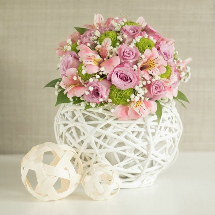 A centerpiece crafted with careful artistry rests atop a smooth white surface, its generous bouquet bursting with harmonious shades of pink, lavender, and sparkling lime green. In this scene-evocative of a refined Queensbury reception or intimate gathering-full roses bloom with velvety softness, flanked by delicate pink alstroemeria adorned with their signature inner streaks and subtle trumpet forms. Spirited clusters of bright, round green button chrysanthemums introduce a playful contrast, their compact forms echoing the joy of spring mornings in local gardens. Wisps of tiny white baby's breath create an aura of gentle airiness, while deep green leaves offer grounding texture among the bright blossoms. Cradling the arrangement is a chic, white vase with an open-weave, lattice-like design-its robust, rounded silhouette a nod to modern style and artisanal craftsmanship, much like the decor seen in Queensbury's evolving homes and event venues. To its left, two decorative spheres in woven neutral tones anchor the scene with an extra flourish-the larger set close to the vase, the smaller poised just ahead. The softly textured beige background ensures the colours sing, creating a sense of freshness and sophistication. This detailed image captures both the beauty of fresh flowers and the spirit of contemporary floral design, making it an ideal muse for local weddings, celebratory centerpieces, or moments shared at home in the Queensbury community.