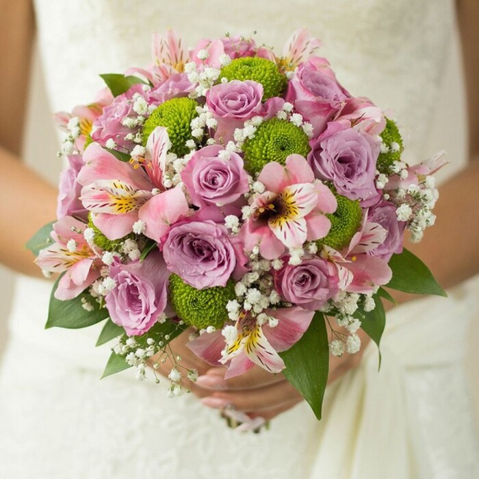 Captured just steps from the leafy avenues of Queensbury, this lush round bridal bouquet rests in the gentle hands of a bride, its vivid color palette echoing the joyful mood of a spring wedding. Crafted with impeccable skill, the bouquet blends soft pink and lavender roses at its heart-some buds tightly furled, others just beginning to open, their silky petals retaining traces of fresh morning dew. Vibrant lime green button chrysanthemums, with their perfect pom-pom shapes, are artfully nestled among the roses, providing a modern and energetic twist on classic elegance. Interspersed throughout the arrangement, pink alstroemeria lilies with trumpet-shaped petals display intricate maroon streaks and cheerful yellow highlights, bringing a unique textural contrast that feels both exotic and inviting. Wisps of white baby's breath-each cluster as light and cloudlike as a breath of fresh air-are woven in, lending softness and a hint of whimsy to the bouquet's volume. Deep, glossy green foliage peeks from beneath, grounding the bouquet and drawing attention to its layered construction. Against the blurred white lace and elegant embroidery of the bride's gown, the bouquet's rich details are rendered in stunning clarity, from the fine veining of the leaves to the interplay of colour and light. In the context of Queensbury's vibrant community celebrations, this bouquet is a glowing symbol of joy and new beginnings, making it an exceptional choice for those seeking wedding flowers that combine classic charm with contemporary style, always freshly prepared for local ceremonies.