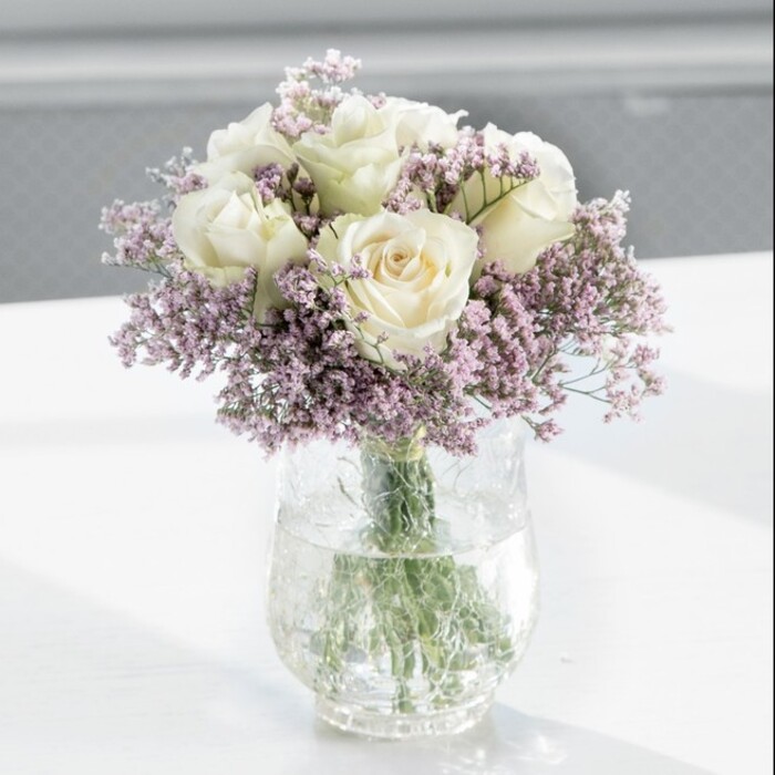 A beautifully composed bouquet of white roses and lilac statice stands at the centre of the scene, crafted with the kind of quiet precision you'd expect from an experienced local florist in Queensbury. The arrangement is full and rounded, with approximately a dozen creamy white roses forming a smooth dome. Each rose is at that perfect stage of openness where the petals spiral outward in soft layers, the outermost ones just starting to relax, giving a plush, almost velvety appearance. Their colour sits between ivory and warm cream, catching the soft daylight and reflecting it gently, so the blooms appear to glow from within. Threaded through and around the roses, fine sprays of lavender-tinted statice create an airy veil, with clusters of tiny, papery flowers on slender stems. These lilac accents add a whisper of colour and an intricate, lace-like texture that contrasts with the more substantial rose petals, giving the bouquet a light, cloud-like halo. All the stems are gathered into a tall, cylindrical clear glass vase, the lower portions submerged in clean water that magnifies the bright green stems as they cross and weave against the glass. The vase stands on a smooth, white surface-likely a table in a bright living room or a reception desk in a small Queensbury office-where the natural light, perhaps streaming in from a nearby window overlooking local streets like Turner Road, is diffused and gentle. The softly blurred background ensures the viewer's focus is fixed solely on the bouquet's serene beauty. The overall impression is calm, refined and timeless, suitable for marking a peaceful celebration, offering condolences with grace, or simply adding a touch of understated luxury to everyday life in Queensbury.