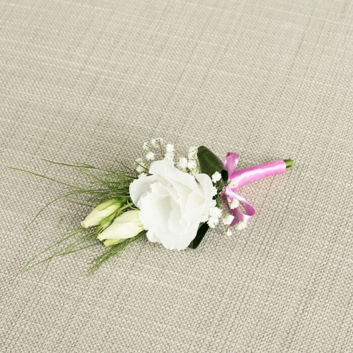 An elegant boutonnière, carefully composed for a wedding or formal event, is photographed from above as it rests at a diagonal angle on a light beige, textured cloth that resembles finely woven linen or burlap. The neutral background, with its subtle criss-cross threads, provides a calm, rustic canvas that highlights every detail of the floral design. At the centre of the piece is a fully opened white eustoma, also known as lisianthus, positioned slightly to the right of the frame. Its petals, soft and gently ruffled, unfold in layered whorls, creating a lush yet delicate focal point that seems almost luminous against the warm fabric. Just to the left of this central bloom sit two slender, unopened eustoma buds in creamy pale green, their smooth, tapered forms adding a sense of natural progression, like the stages of a relationship growing over time-from first dates in Queensbury's local pubs to the moment of exchanging vows. Fine sprays of tiny white baby's breath flowers are interwoven among the eustoma blooms, their fragile, cloud-like clusters giving the boutonnière lightness and an ethereal charm familiar in classic wedding florals. From behind the main flowers, narrow, dark green, grass-like leaves stretch gracefully toward the bottom left corner, adding movement and a fresh, garden-picked feel. A broader, darker green leaf is tucked subtly behind the white bloom, grounding the composition and offering contrast. At the lower right, the stems are gathered neatly and wrapped in a vivid fuchsia satin ribbon, tied into a small, lively bow. This bright pink accent introduces a modern, playful note against the timeless white and green palette, much like the colourful touches often chosen by couples planning weddings around Queensbury. Crafted with the care of an experienced florist, the boutonnière appears ready to be pinned to a lapel, its blend of textures, tones, and shapes conveying quiet joy and refined celebration.