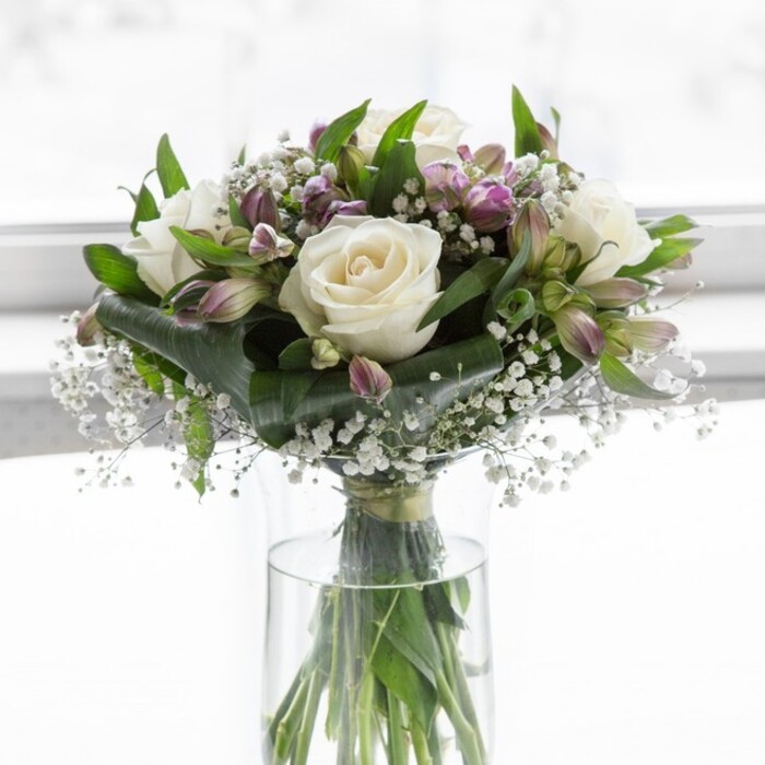 A tall, cylindrical glass vase stands at the centre of the scene, showcasing a lush, rounded bouquet composed of creamy white roses, lavender-toned alstroemeria, and soft clouds of baby's breath, all bathed in diffused daylight. The roses are fully open yet still pristine, their silky petals layered in gentle spirals that create a plush, almost cushion-like texture. Surrounding and weaving around them, the alstroemeria introduce notes of cool lilac and muted purple; each stem bears both tight, green-held buds and freshly opening blooms, their narrow petals carrying fine, darker streaks that draw the eye inward. Fine, branching stems of gypsophila thread throughout, adding hundreds of tiny white blossoms that fill gaps, soften edges, and lend the bouquet a dreamy, floating quality. Deep green foliage forms a natural frame: broad, glossy leaves cluster closer to the vase, while more slender leaves and stems rise to support individual flowers, giving the arrangement structure and a sense of movement. Through the clear glass, the neatly trimmed stems are visible in transparent water, angled with precision that speaks of careful, professional floristry. The background is pale and softly blurred, perhaps a bright wall or window in a Queensbury townhouse, so the flowers appear to glow against their surroundings. The overall feeling is one of calm refinement and heartfelt sincerity, suited to moments when words might feel too heavy, such as offering comfort after a loss, marking an anniversary, or wishing someone quiet strength. It's easy to imagine this bouquet being hand-delivered to a home off Honeypot Lane or near Queensbury Underground, arriving as a gentle, fragrant reminder of care and presence.