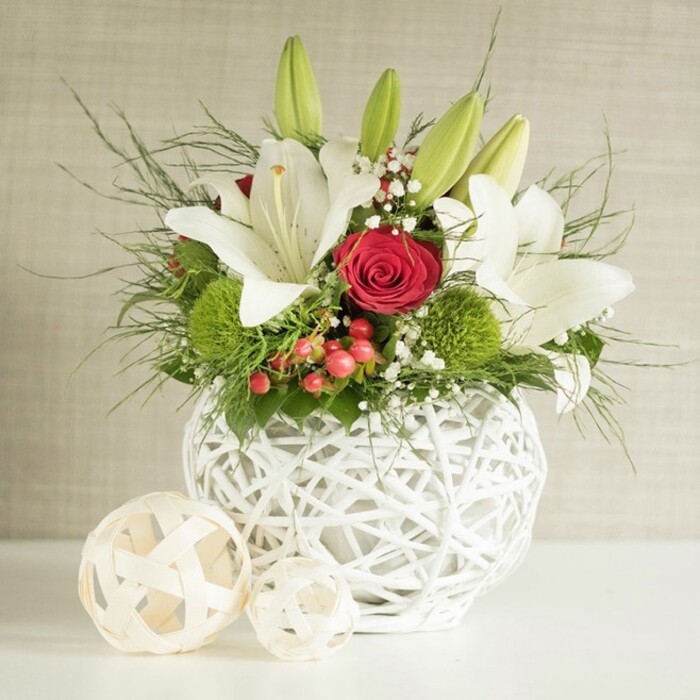 This image shows a beautifully composed floral display that could easily sit at the centre of a wedding table in Queensbury, catching the eye of guests as they settle in for the reception. The arrangement is presented in a round, bright white vase constructed from interwoven strands, creating a basket-like, open-weave pattern that feels both sculptural and light. From this textured vessel rises a bouquet rich in contrast and detail. Several large white lilies arch gracefully outward, their petals wide and silky, revealing faint speckles and warm yellow stamens that suggest a gentle, sweet fragrance. Some lilies are already in full bloom, while others remain in tapered green bud form, adding a sense of freshness and unfolding time to the scene. Interspersed among the lilies are lush, velvety red roses, one especially prominent bloom positioned slightly off-centre, its tightly furled spiral of petals offering a classic symbol of romance. To break up the smoothness of these focal flowers, bright green, spherical pompons-likely dianthus or chrysanthemums-are tucked into the arrangement, their dense, cushiony texture introducing a fun, modern twist that would appeal to couples marrying in the Queensbury area. Fine, delicate clusters of white gypsophila weave through the flowers like a soft mist, while small sprays of red berries provide additional points of colour that hint at festive celebrations or winter weddings. Around and beneath the blooms, a mixture of greenery-including slender, feathery fronds and deeper, broader leaves-gives fullness and natural movement. On the white surface beside the vase, two decorative orbs crafted from pale woven material mirror the vase's texture and create a cohesive, stylish vignette. A softly textured neutral wall behind the arrangement keeps attention firmly on the interplay of white, red, and green, capturing the mood of quiet sophistication and heartfelt celebration.