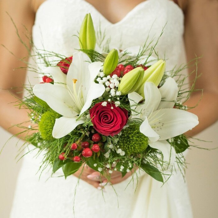 A detailed close-up brings into focus a local Queensbury bride, her fingers gently curved around an exuberant bridal bouquet positioned against the fine white bodice of her textured strapless dress-soft ripples of fabric echoed by each petal. The composition exudes classic charm, with multiple stately white oriental lilies, some in glorious full bloom displaying delicate golden stamens, while others remain as fresh, green-tipped buds ready to blossom-capturing moments of both beauty and promise. At the centre, a deep crimson rose stands as a symbol of love and romance, surrounded by whispers of white baby's breath and cheerful red hypericum berries-a touch inspired by walks along Honeypot Lane. Green button-like clusters, resembling miniature moss patches or green trick dianthus, peek playfully from among the stems, tying together the wedding's lively palette. Cascades of feathery fern fronds and broader leaves are interwoven, giving the bouquet a natural fullness reminiscent of arrangements crafted by experienced hands at Flower Delivery Queensbury. The dreamy, softly blurred backdrop allows the vibrant floral details to shine, making this an exemplary choice for both traditional and celebratory weddings in Northwest London.