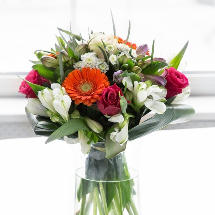 This radiant bouquet, meticulously crafted in the heart of Queensbury, is a vibrant homage to natural beauty and community spirit. At its heart, joyful gerbera daisies in hues of sunset orange and gentle peach spread their petals wide, their velvety textures catching the play of natural light just as blossoms do on a sunlit morning by Queensbury Circle. Deep magenta roses add passionate, spiralling depth, nestled beside airy sprays of pristine white alstroemeria which lend lightness and a subtle, understated grace. Here and there, small white and pale pink blooms emerge, their delicate textures adding layers of complexity and surprise, much like the patchwork gardens hidden throughout the local neighbourhood. Encasing this bouquet, glossy green aspidistra leaves are elegantly folded, creating a structured collar at the base-a nod to the reliable, enduring greenery seen across Queensbury's open spaces. The slender, crystal vase reveals a tangle of vibrant stems, each carefully aligned, highlighting the artistry and precision behind the arrangement. Soft, diffuse light lends each flower enhanced vibrancy while retaining a gentle sense of calm. This ensemble evokes the scent of a spring morning-sweet, fresh, and quietly luxurious. Perfect for a joyful celebration, a thoughtful thank you, or to simply infuse a local home with optimism, this bouquet reflects the attentive care of an expert florist and the lively heart of Queensbury.
