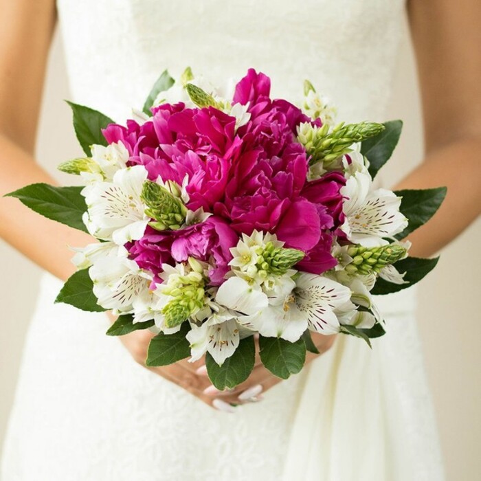 In this intimate wedding photograph, the camera focuses tightly on a bride's midsection as she holds a round, hand-tied bouquet, the flowers resting gently against the soft white fabric of her gown. At the centre of the arrangement, large, deeply ruffled peonies in a vivid fuchsia-magenta shade create a dense core of colour, each petal overlapping like folds of velvet, full and slightly cupped as though caught at the peak of bloom. These opulent blossoms are surrounded by pure white alstroemeria, their long, slender petals opening outward and decorated with fine maroon speckles deep inside, adding an intricate pattern that rewards a closer look. Nestled throughout the bouquet are unusual spiky green buds and starry filler flowers, their upright, pointed shapes bringing a modern, textural contrast to the rounded softness of the peonies. Around the outside, broad, glossy green leaves frame the design, their darker tones grounding the brighter colours and giving the bouquet a natural, garden-inspired finish reminiscent of floral displays seen around Queensbury and the nearby green spaces. The bride's hands are clearly visible at the bottom of the image, fingers interlaced around the stems, nails neatly manicured in a delicate pale pink that echoes the romantic palette. The background is softly blurred, leaving only a hint of lace detail from the dress, so that the viewer's attention remains entirely on the flowers and their freshness. It feels like a bouquet created for a joyful ceremony at a local Queensbury venue, carefully composed by a florist who understands how to blend classic bridal elegance with a touch of contemporary flair, ideal for late spring or summer celebrations.