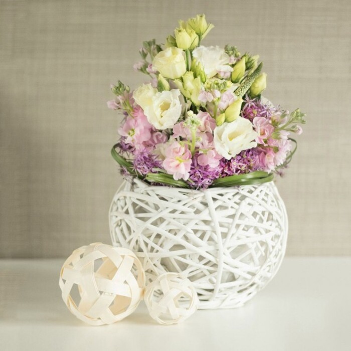 A tranquil bouquet, styled by an expert Queensbury florist, occupies the center of a crisp white tabletop, its colours and textures evoking the quiet elegance of spring mornings across Brent's leafy boulevards. The arrangement overflows with creamy eustoma, each flower's ruffled petals tinted with the subtlest shades of yellow and white, creating a plush visual softness. Sprigs of pink stock flowers contribute gentle verticality and a whisper of fragrance, while the underlay of petite purple fillers-mini asters or allium-echoes the vibrant, natural palette found in local community gardens. The composition is thoughtfully supported by a wide border of deep green foliage that both frames and enhances each bloom. Housed in a distinctive, open-work white vase-its spherical shape fashioned from intricately woven rattan or painted twigs-the bouquet achieves a balance between airy modernity and organic abundance. To the left, two decorative spheres, crafted in matching rattan open-weave, complete the vignette, their presence nodding to the handmade décor traditions of the area. The softly textured beige background, suggestive of elegant local interiors, gently illuminates the florals, casting a serene glow that captures Queensbury's blend of tradition and modernity. This envisioning is suited for everything from wedding table arrangements to special dinner celebrations in and around Queensbury.