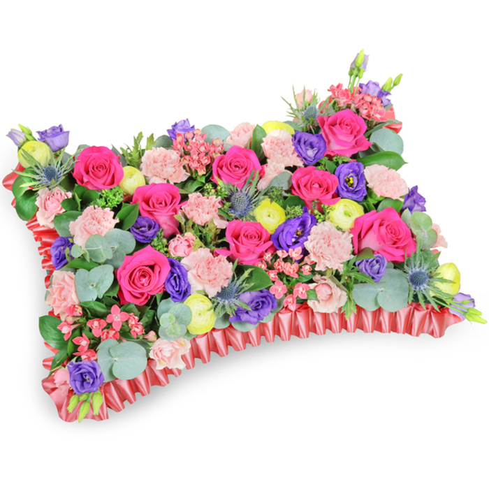 Captured from a slightly elevated angle against a pure white field, this pillow-style memorial arrangement is a study in harmonious contrasts and sympathetic craftsmanship. A sea of hot pink roses spreads across the top, their satiny petals forming lush, tactile islands that draw the eye inward. Interwoven are soft blush carnations, whose ruffled masses create gentle transitions between rose clusters and other accents. Deep purple lisianthus punctuate the composition with cool, ceramic-like tones, while lemon-yellow ranunculus provide bright, optimistic highlights-small, layered suns that lift the palette toward celebration. Spear-like, silver-blue eryngium thistles add a bracing, architectural note; their spiky forms break up the roundness and lend an unexpected note of wild beauty. Rounded eucalyptus leaves, bluish-silver in the light, trail through the arrangement and offer a subtle evergreen scent that calms the overall impression. The flowers sit neatly enclosed by a pleated red satin or silk ribbon base, the ruffled edge forming a defined rectangular cushion and providing an elegant frame. Texturally, the piece alternates velvety, frilled, and spiky elements so that each glance finds a new detail; structurally, it is densely wired and backed to hold its pillow silhouette at services. Often chosen for wakes or graveside remembrances around Queensbury station, the local crematorium or for floral tributes delivered to families on Chalkhill Road, this arrangement reads as a comforting celebration of a life: colourful, tender, and thoughtfully composed by an experienced neighbourhood florist.