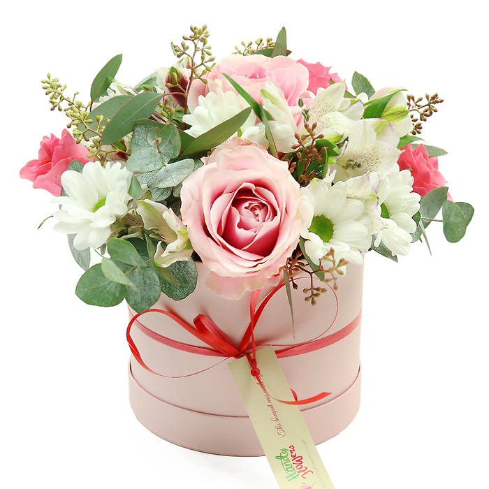 Set against a bright white background, a chic pastel pink hatbox brims with an abundant floral display, the entire scene composed with the precision of an experienced Queensbury florist. The box is perfectly cylindrical with a velvety matte surface, its gentle blush tone giving it a soft, romantic character. Around the base, a narrow coral-red satin ribbon is wrapped cleanly and tied into a small but confident bow at the centre front. Suspended from this bow hangs a rectangular cream tag with elegant green, handwritten-style script, evoking the personal touch of a note tucked in before the bouquet is sent out for local delivery. Overflowing from the top, the flowers form a generous, domed arrangement. Several light pink roses, some touched with deeper fuchsia at their cores, open luxuriantly, their layered petals creating a focal point that suggests tenderness and celebration. These are interspersed with neat white chrysanthemums or daisy-like blooms, each with a vivid yellow-green centre that brightens the whole palette and recalls the fresh greens seen in neighbourhood gardens around Queensbury. Slender stems of white alstroemeria, with their graceful trumpet shapes and subtle inner streaks, weave elegantly between the rounder blossoms, adding height and finesse. Deeper pink blooms-possibly small roses or lisianthus-pop here and there, enriching the mix with more intense colour and giving a sense of joyful contrast. Sprays of eucalyptus in soft, silvery green extend beyond the main flowers, joined by slimmer dark green leaves and delicate reddish-brown buds or seed pods that lend a slightly rustic, organic quality. Together, these elements create a refined yet approachable hatbox arrangement, ideal for anniversaries, Mother's Day surprises, or thoughtful get-well wishes delivered anywhere across Queensbury, capturing both modern style and heartfelt sentiment.