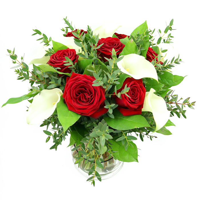 An artfully composed bouquet of red roses and white calla lilies sits centred in a clear glass vase, set against a clean white background that heightens its rich colours and refined shape. Around six or seven red roses, fully bloomed and luxuriously velvety, anchor the arrangement, each blossom showing layers of deep crimson petals that curl inward to form intricate spirals. These classic roses are interspersed with four or five pristine calla lilies, their long, tapered stems and trumpet-like blooms giving the design a modern, sculptural feel. The calla petals are a creamy, almost porcelain white, folding around slim yellowish-orange centres and adding a cool contrast to the warm red of the roses. Surrounding the blooms, generous sprays of greenery create texture and volume: slender eucalyptus stems with small, rounded, silvery-green leaves weave through the flowers, lending a slight, windswept softness, while broader, darker emerald leaves build a solid, glossy base that feels both natural and elegant. The cylindrical vase reveals the gathered stems and clear water inside, with a delicate ribbed detail close to the base catching the light and adding subtle visual interest. Bright, even illumination brings out the freshness of each petal and leaf, making the arrangement appear as though it has just left a Queensbury florist's workbench. This bouquet would sit beautifully on a dining table in a home near Queensbury Tube station or brighten an office along Honeypot Lane, ideal for anniversaries, romantic surprises, or any occasion where a polished, heartfelt floral gesture is needed.