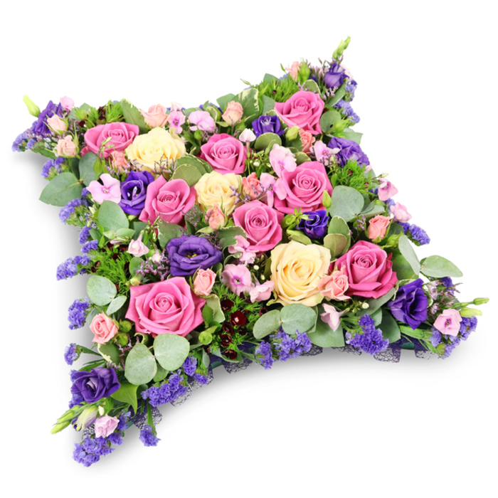 A richly detailed funeral cushion in the shape of a soft, rectangular pillow is shown against a crisp white background, a faint shadow underneath giving the impression that it has just been set down with quiet reverence. The entire surface is completely covered in flowers, forming a dense, velvety layer of colour and texture. At the centre and radiating outward, generous-headed roses in vibrant fuchsia pink, tender blush, and creamy pale yellow or ivory open in full, their petals curling back to reveal perfect rosettes. Interwoven among these are elegant deep purple lisianthus, their gently ruffled blooms adding a note of formality and depth, almost like satin ribbons threaded through a tapestry. Smaller pink spray roses and tiny, cloud-like clusters of lavender and mauve filler flowers, possibly statice or limonium, fill the spaces between the larger stems so that no foliage or base is visible from above. Along the edges and tucked throughout, rounded, silvery-green eucalyptus leaves soften the palette, while darker, ferny greenery slips between the petals, creating contrast and a sense of natural movement. The overall cushion has rounded corners and a softly domed profile, resembling a plush pillow that symbolically offers eternal rest and comfort. In the stillness of the white background, the colours appear especially vivid, from the bright berry pinks to the soothing greens and cool purples. It is easy to imagine this tribute prepared by an experienced florist in Queensbury, ready to be delivered to a local chapel, crematorium, or cemetery, perhaps for a family gathering after a service near the quiet residential roads around Honeypot Lane. This floral cushion conveys dignity, tenderness, and enduring remembrance for someone deeply missed.