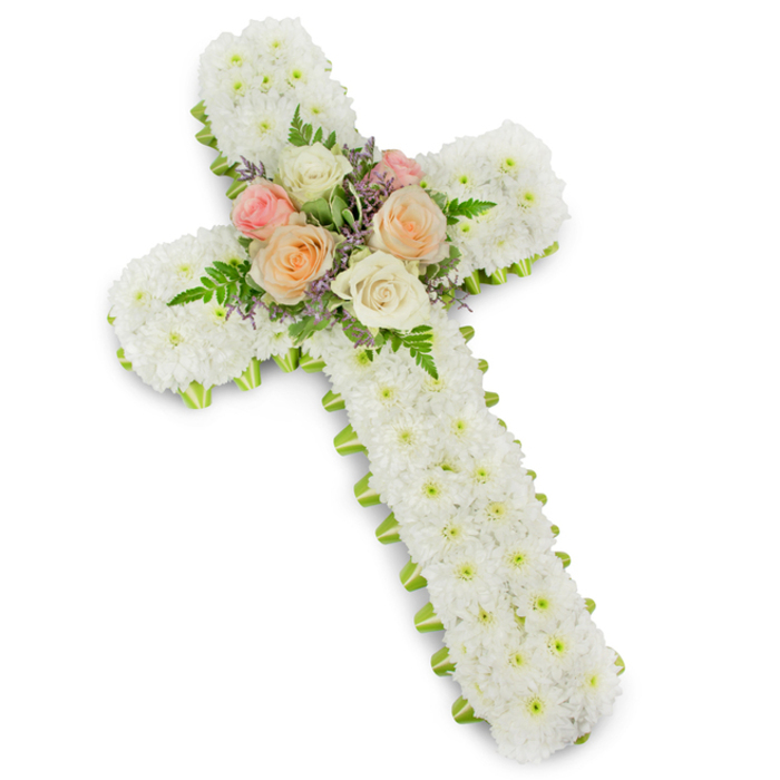 This image presents an elegant Christian cross funeral tribute, perfectly centred against a crisp white background that enhances every detail of the floral work. The structure of the cross is robust and three-dimensional, its entire surface densely carpeted with tiny white chrysanthemum button blooms. Each floret is nestled snugly against its neighbour so that the cross appears as a single, velvety expanse of white, smooth yet richly textured. The symbolism of purity, peace, and spiritual light is immediately apparent in this uninterrupted sea of white petals. Around the perimeter of the cross runs a precise band of lime green ribbon, subtly visible beneath the edge of the flowers. This slim border of fresh green creates a clean outline, giving definition to the cross's contours and adding a hint of contemporary brightness without disturbing the design's solemn nature. At the intersection of the arms and vertical beam, a thoughtfully composed cluster of focal flowers draws the eye. Several fully opened roses in shades of ivory, muted blush pink, and soft peach form a gentle heart at the centre. Their larger petals and spiralling forms bring softness and grace to the composition. Threaded among them are delicate fronds of green fern and airy sprays of lavender or light purple filler blossoms, adding texture, movement, and a whisper of colour contrast. The mood is contemplative and reverent, evoking the quiet stillness of a farewell service at a Queensbury chapel or a memorial gathering near the open spaces of nearby parks. This type of floral cross, often ordered for funerals or graveside tributes, reflects the skilled, patient hands of a local florist who understands how flowers can express faith, remembrance, and enduring affection when words are hard to find.