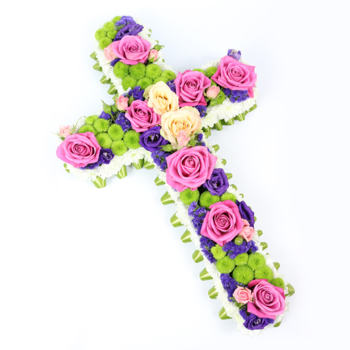 Viewed from above against a crisp white background, this meticulously crafted floral cross dominates the image with its balance of structure and softness. The silhouette is clean and unmistakable, thanks to a thick edging of tiny white chrysanthemums that run continuously along every curve and corner, forming a delicate, cloud-like frame that evokes purity and peace. Just beneath this white fringe, a narrow band of carefully folded light green ribbon or foliage traces the same path, like a discreet border of fresh grass or new leaves, giving the design a subtle two-tone outline that feels polished and intentional. Inside this border lies a rich mosaic of flowers. Large roses in shades of hot pink and creamy pale yellow are distributed along the central stem and out across the arms of the cross, their generous blossoms opening fully to reveal layers of velvety petals. Their colours alternate naturally, catching the light differently and providing gentle highlights and shadows. Clustered among these roses are lively lime-green button chrysanthemums, their compact, rounded heads forming bright accents that punctuate the arrangement, almost like beads or jewels set between the larger blooms. Smaller flowers in deep purple and violet, probably lisianthus or statice, weave through the gaps, filling the spaces with colour and giving the design depth from every angle. The overall lighting is bright yet soft, with no harsh shadows, allowing the viewer to appreciate every nuance. This floral tribute would feel at home in a quiet chapel service in Queensbury, perhaps near the green spaces around Queensbury Park, offering a comforting presence that speaks of faith, remembrance, and the enduring beauty of a life remembered. Flower Delivery Queensbury could present such a cross for same-day delivery to local funerals, ensuring it arrives as fresh and serene as it appears here.