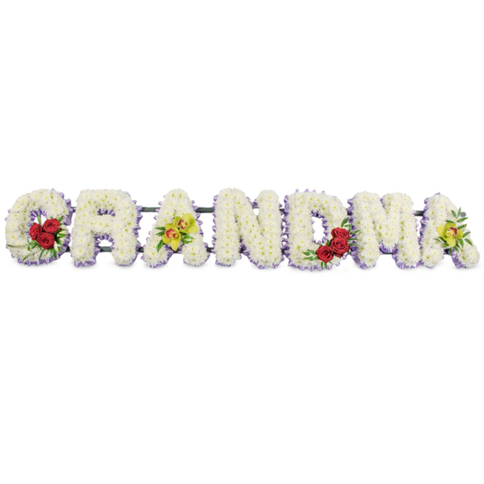 A dignified floral tribute spelling GRANDMA stretches horizontally across a plain white background, as if prepared for a memorial display in a peaceful Queensbury chapel or family gathering. Each capital letter is sculpted from a thick bed of small white chrysanthemums, their rounded petals overlapping to create a cushion-like surface that feels both soft and substantial. The flowers are packed so closely that the letters appear almost carved from fresh blossoms, with no bare spaces, giving the tribute a polished, professional finish. Just beneath the white blooms, a gentle trim of light lavender petals traces the base and edges of each letter, forming a delicate outline that adds definition without overwhelming the design. Carefully chosen accent flowers are placed at significant points to convey intimacy and personal meaning. On the opening G, three deep red roses cluster on the lower left, their rich colour and velvety texture speaking of enduring love and respect. The first A features two yellow-green orchid blooms positioned gracefully along its central bar, their reddish-brown patterned throats adding a hint of exotic detail that might recall a favourite corsage or gift. The R, N, and D remain simple and pure, composed solely of the white flowers and lavender edging, providing a sense of stillness in the middle of the word. Towards the end, the M carries another trio of red roses in its lower right arch, while the final A once again displays two luminous chartreuse orchids, echoing the earlier motif. A dark green metal frame discreetly supports each letter from behind, allowing the entire tribute to be placed on a stand or hearse. The overall look is serene, traditional, and deeply affectionate-a handcrafted farewell from family, reflecting the quiet strength and tenderness of a much-loved grandmother.