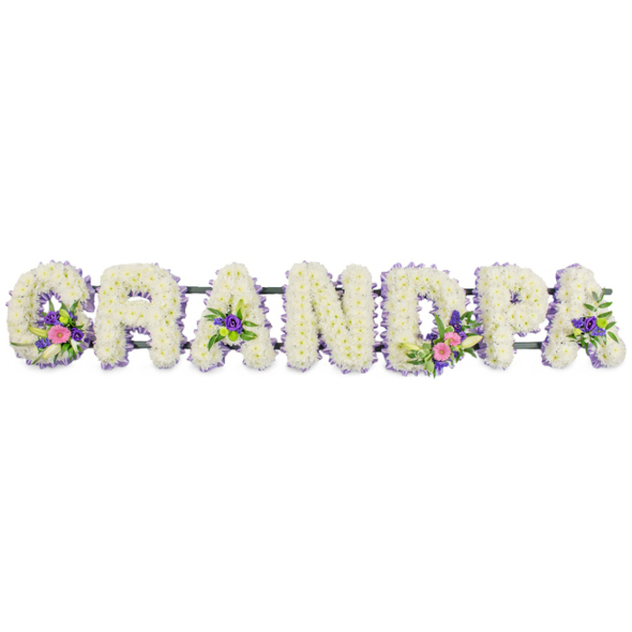 The image shows a personalised funeral flower display spelling GRANDPA in large, upright letters, created as a loving tribute for a beloved grandfather. Each letter is solidly constructed from masses of small white flowers-most likely chrysanthemums or spray carnations-so tightly packed that they form a smooth, plush blanket of petals, like soft snow resting in even layers. Around the contours of every letter runs a slim edging of tiny purple and lilac blooms, perhaps statice, asters, or heather, lending a gentle contrast and defining the shapes with a cool, lavender halo. Within the pristine white base, the florist has worked in clusters of brighter accent flowers: the 'G' holds a small spray of pink or fuchsia blossoms, a refined white lily-style flower, and glossy green foliage; the first 'A' echoes this with a subtle grouping of purple blooms and greenery. The 'D' carries a more prominent cluster that mirrors the 'G', again combining bright pinks, white lily petals, and fresh green leaves, while the final 'A' is finished with rich purple florets and dark foliage. All seven letters are fixed to a low, dark green frame running horizontally behind, designed so the tribute can stand securely at a service, perhaps in a Queensbury church or community hall. The plain white background of the photograph, with its soft, even lighting, focuses attention entirely on the flowers and their craftsmanship. The arrangement feels both contemporary and traditional, the kind of bespoke sympathy tribute a family in Queensbury might choose to express their love, memories, and respect for Grandpa in a way that words alone cannot.