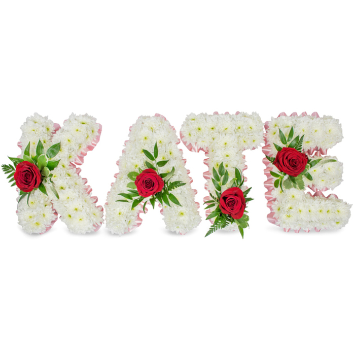 A dignified, handcrafted floral arrangement spells the name KATE in substantial block lettering, photographed against a crisp white field so every floral detail is clear. Each letter is formed from a voluminous mass of pristine white chrysanthemums, their compact, cushiony heads arranged to create smooth, cloud-like surfaces that convey purity and solemn grace. The outer contours are trimmed in a pale pink satin ribbon, the gentle pleat and luster of the ribbon providing a soft, elegant frame that offsets the chrysanthemum's matte texture. At the heart of each letter a solitary red rose provides a vivid, emotive counterpoint: the roses' velvety, deep crimson petals sit proudly amid the white, signalling love and enduring memory. Complementary green foliage - glossy laurel-like leaves with finer fern accents - surrounds each rose, adding shadow, depth and a feeling of renewal. Thoughtful placement of the roses enhances legibility and balance: the K's floral accent is positioned on the lower part of its left upright; the A's rose marks the peak of its triangle; the T's bloom rests at the left base of its crossbar; and the E's rose highlights the centre horizontal bar. The overall effect is a classic sympathy tribute, both visually clean and emotionally resonant, suitable for funeral chapels or memorial services in Queensbury and neighbouring areas such as Burnt Oak and Colindale. A local florist's touch is evident in the careful textures, scent profile and considered colour contrasts.
