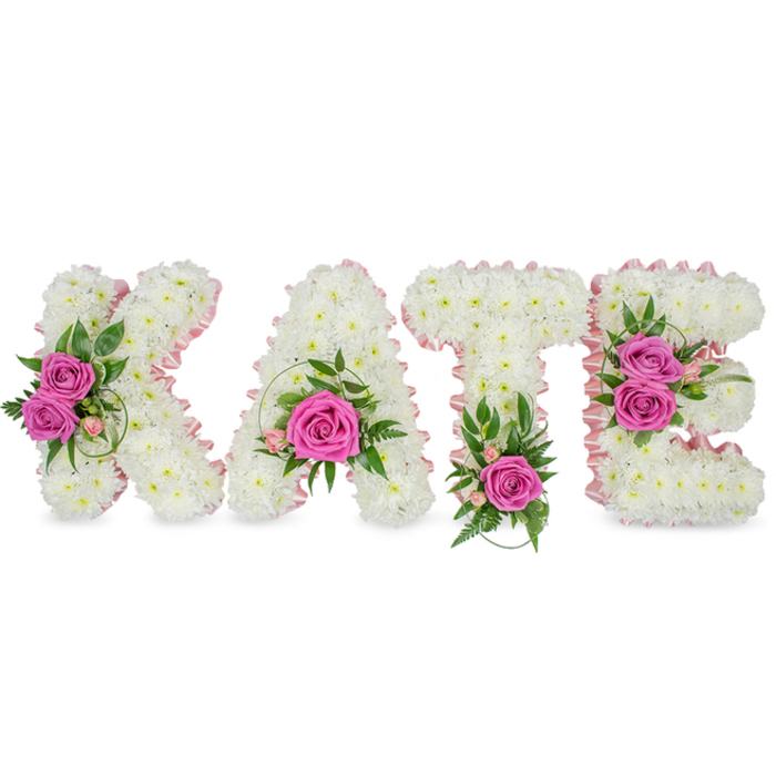 A striking and intimate memorial piece spells out the name KATE in voluminous, block-style letters crafted entirely from hundreds of small, immaculate white chrysanthemums. Each chrysanthemum is placed closely to create a smooth, cloud-like surface that invites the eye to travel across its plush texture, while a narrow pale pink satin ribbon outlines the inner shapes of every letter, giving a soft, feminine edge that shimmers faintly under soft lighting. Vivid hot pink roses are used sparingly and deliberately as focal gems: two roses sit amid the left arm of the K, a single bold rose rests at the base of the A framed by delicate pink buds and sprigs, a lone rose anchors the bottom of the T accompanied by fern-like foliage, and two roses brighten the interior curve of the E, enhanced by variegated leaves and wisps of greenery. The composition rests freestanding against a pristine white backdrop with subtle shadowing that emphasizes its three-dimensional form - suitable for placement beside a casket or on a remembrance table. The scent is quietly floral, the touch plush and structured; the roses' silkiness contrasts with the chrysanthemums' matte, almost fabric-like appearance. Local residents around Queensbury High Street and the Jubilee line station often select this design for funeral services and commemorations because its clear lettering offers a personal, readable tribute that balances dignity with heartfelt warmth and skilled craftsmanship.