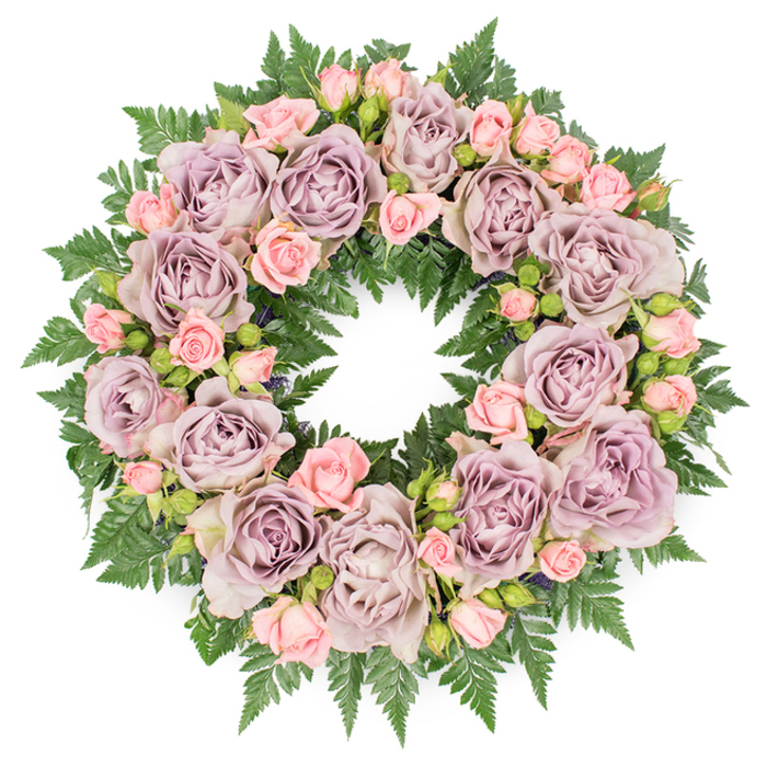 A meticulously arranged floral wreath rests flat on a clean white surface, its circular form perfectly defined by an abundance of roses and fresh greenery. The focal blooms are large, full-faced roses in an elegant palette of dusty lavender, soft mauve, and grey-lavender tones, each one fully open to reveal its intricate, spiralling petals. These roses are evenly spaced around the ring, creating a sense of balance and calm that suits a thoughtful funeral or memorial tribute. Nestled among them are sprays of smaller, blush-pink roses, some as tiny, tight buds, others just beginning to unfurl. This mix of stages brings gentle texture and a feeling of quiet life continuing, an especially comforting detail for families gathering in Queensbury to say goodbye, perhaps after a walk through nearby parks or a service in the local area. Underpinning the pastel blooms is a dense collar of deep green fern fronds, likely leatherleaf or sword fern, with their fine, serrated segments radiating outward to support and frame the flowers. The greenery forms a rich, verdant cushion, its darker tones contrasting beautifully with the delicate pink and lavender roses, making each blossom stand out clearly against the white background. Subtle touches of darker filler foliage and tiny buds are woven discreetly between the roses, adding depth and preventing the design from feeling flat. The lighting is even, without harsh shadows, so every petal and leaf is clearly defined, emphasizing the skill of the florist who assembled it. The wreath conveys a mood of gentle dignity and remembrance, ideal for a final farewell, a celebration of life, or a quiet commemoration within the Queensbury community.